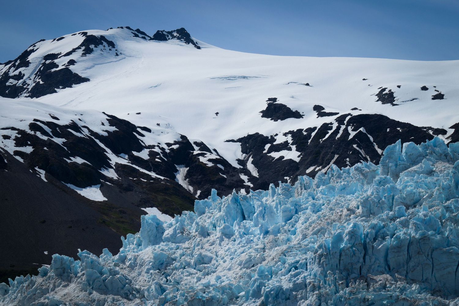 A tidewater glacier in the foreground with jagged glacial ice, and a large snow-draped mountain ridge in the background with blue sky.