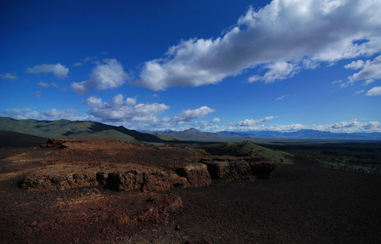 A wide angle photo from near Craters of the Moon National Monument that shows the edge of a massive lava field with mountains in the back.