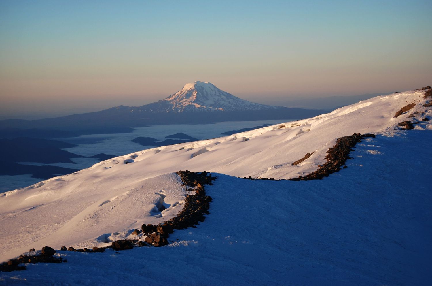 A sunrise view of Mt. Adams from the summit of Mount Rainier, with a ridge of narrow rock that lines one of the summit craters.