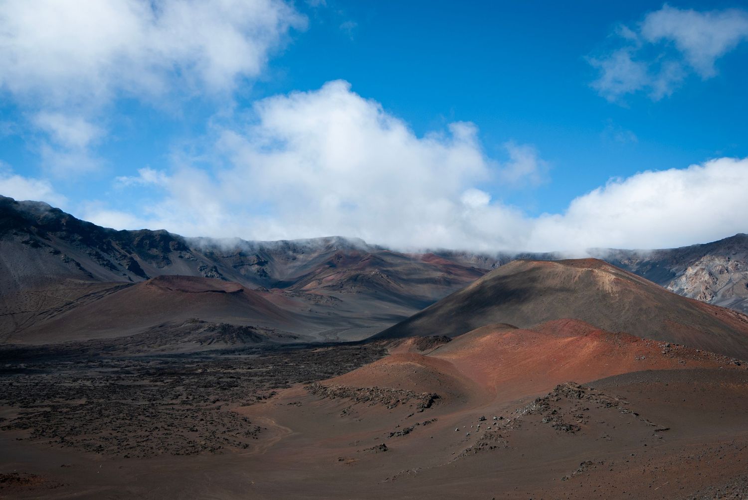 A view inside the crater area of Haleakala, on the island of Maui. Red cindercones and cliffs beneath a cloudy blue sky.