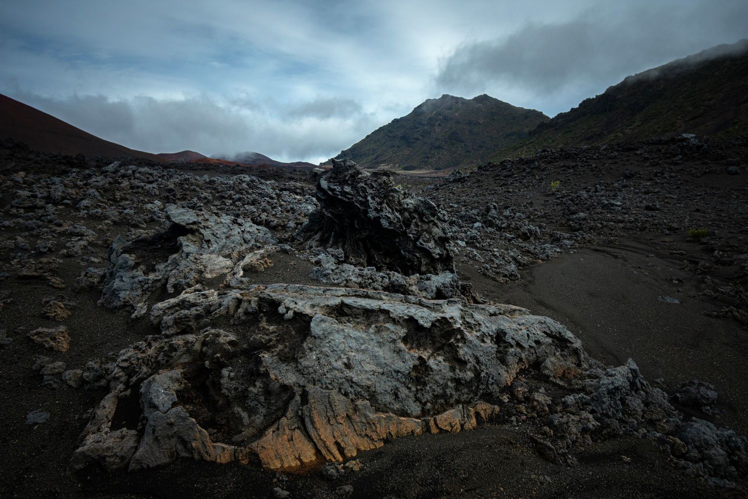 A moody cloudy view of the Mars-like crater area of Haleakala on Maui. Jagged rocks sit in dark volcanic sand.
