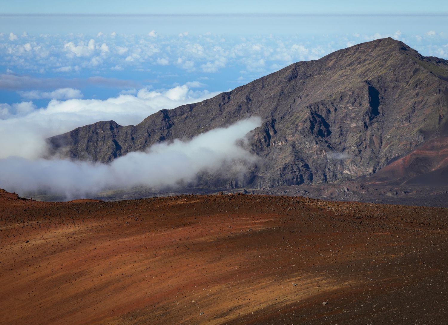 A bank of cloud creeps up a huge peak at the edge of the crater of Haleakala, a 10,000 foot mountain on Maui. The ocean and sky lies beyond.