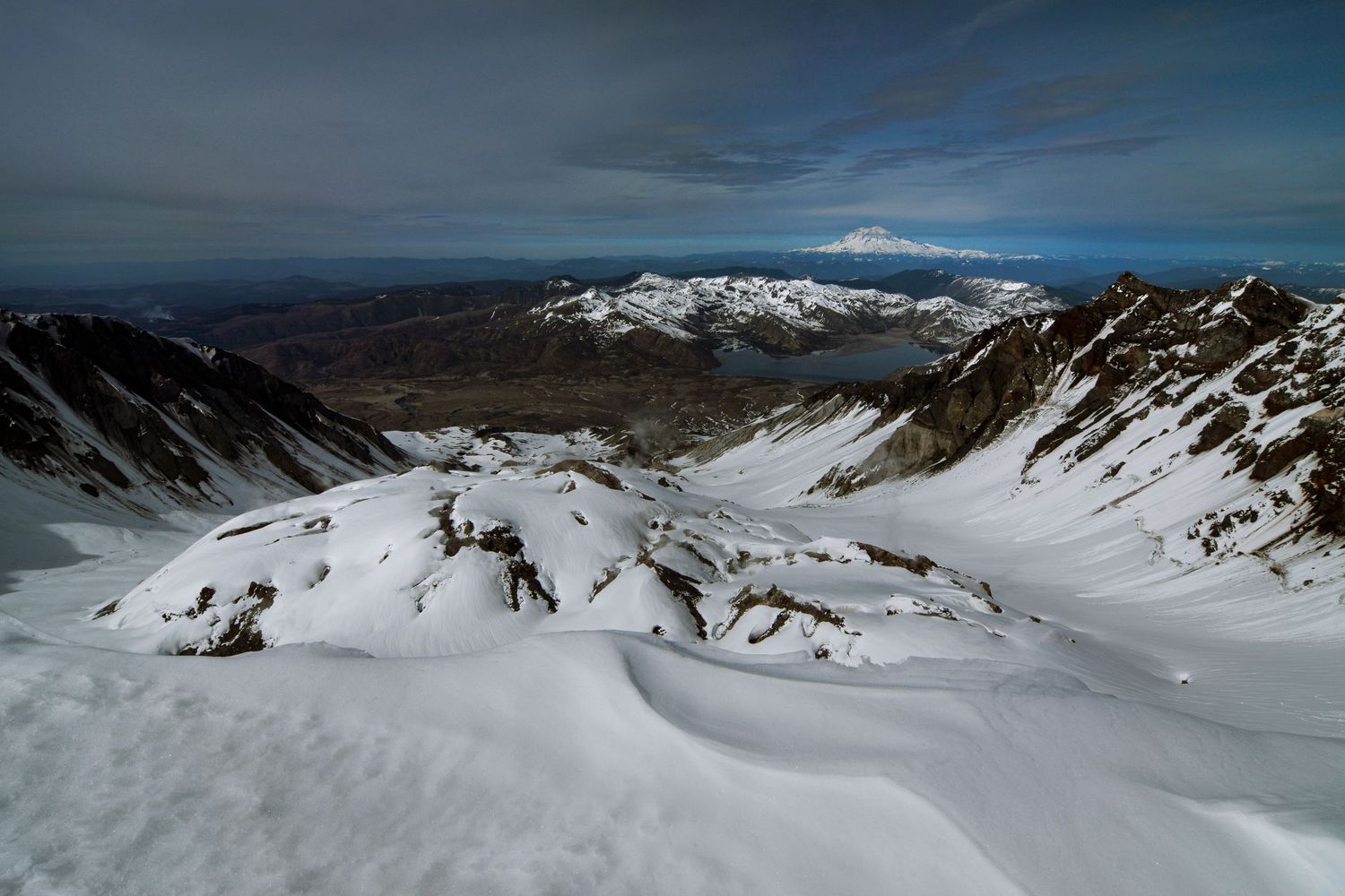 A view from the summit of Loowit or Mount St. Helens in early spring looking down at the snow-covered cone building in its crater.