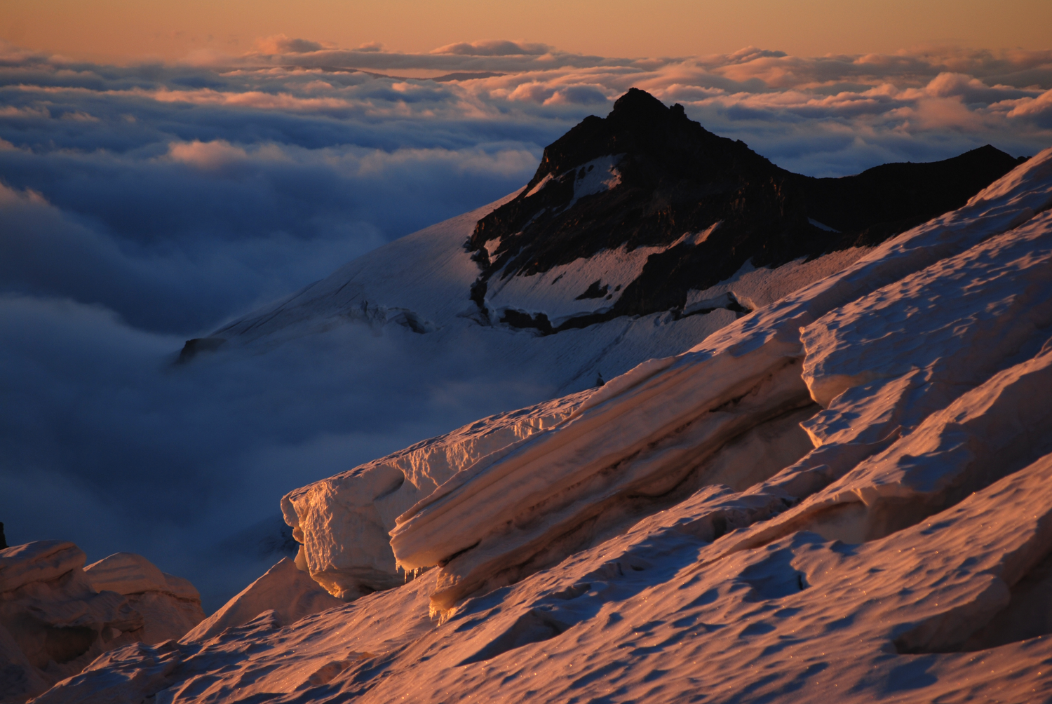 A dramatic sunrise view of sunlit glacier and a peak called Little Tahoma standing above a layered sea of clouds on Mount Rainier.