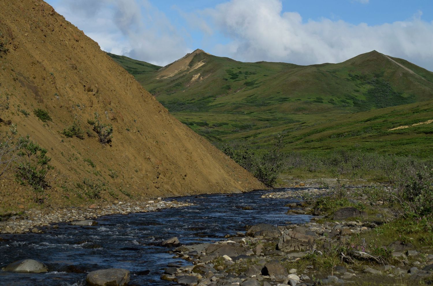 A metallic blue river cuts past a hill of brown soil with verdant green tundra hills on the other side and in the distance.
