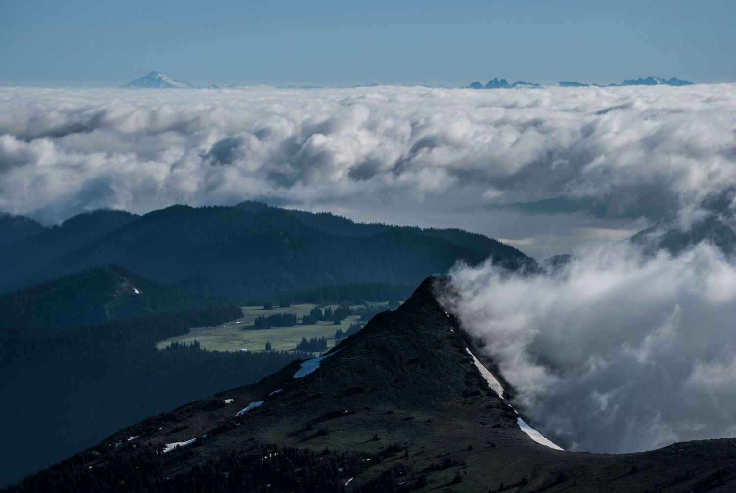 A view of ridges and peaks in Mount Rainier National Park as well as a large sub alpine meadow, with thick clouds pooling all around.