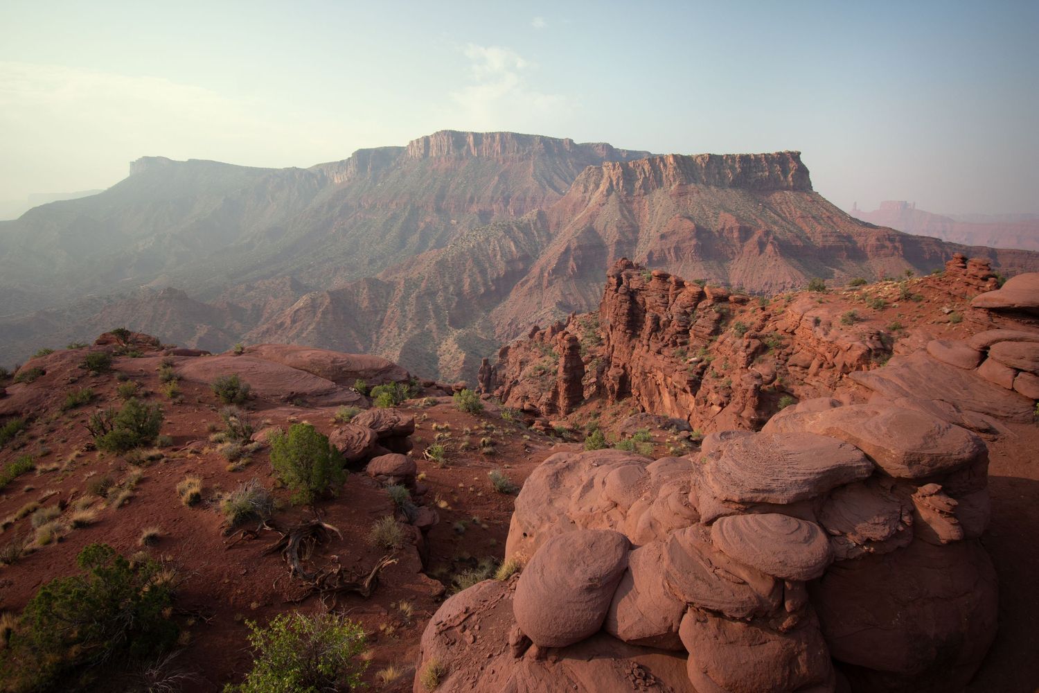 Sandstone cliffs with heavy erosion in the distance and lumpy sandstone bedrock next to red soil with scrub in the Fisher Towers area.