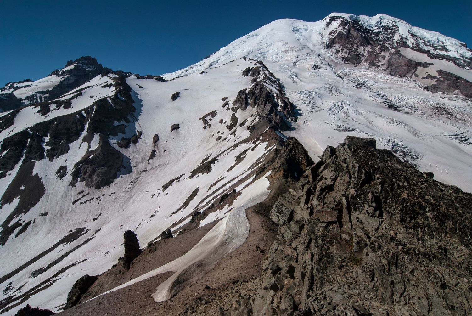 A bright midday view of the north side of Mount Rainier or Tahoma from Third Burroughs; glaciers extend to either side of a rocky ridge.