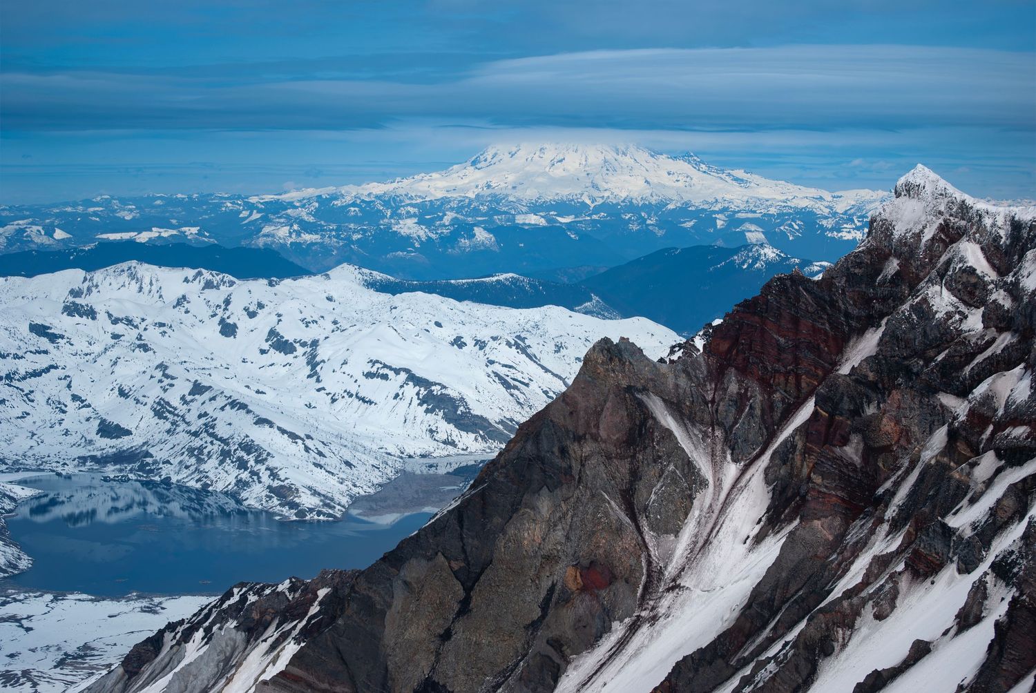A view with one of the insides of Mount St. Helen's crater visible, looking out towards Mount Rainier in the distance, in snowy spring.