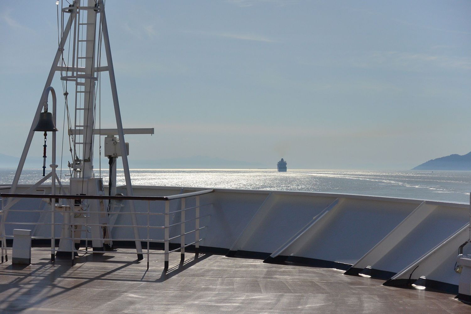 View from the front of a cruise ship looking out onto the sunny ocean with another cruise ship off the in the distance, amid bright waters.