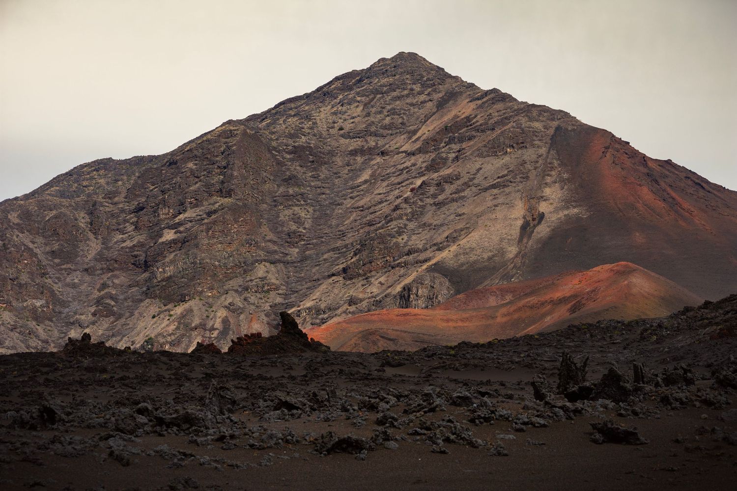 A view of a peak within Haleakala National Park on Maui, red minerals stain the side of the peak and a Mars like foreground sits below.