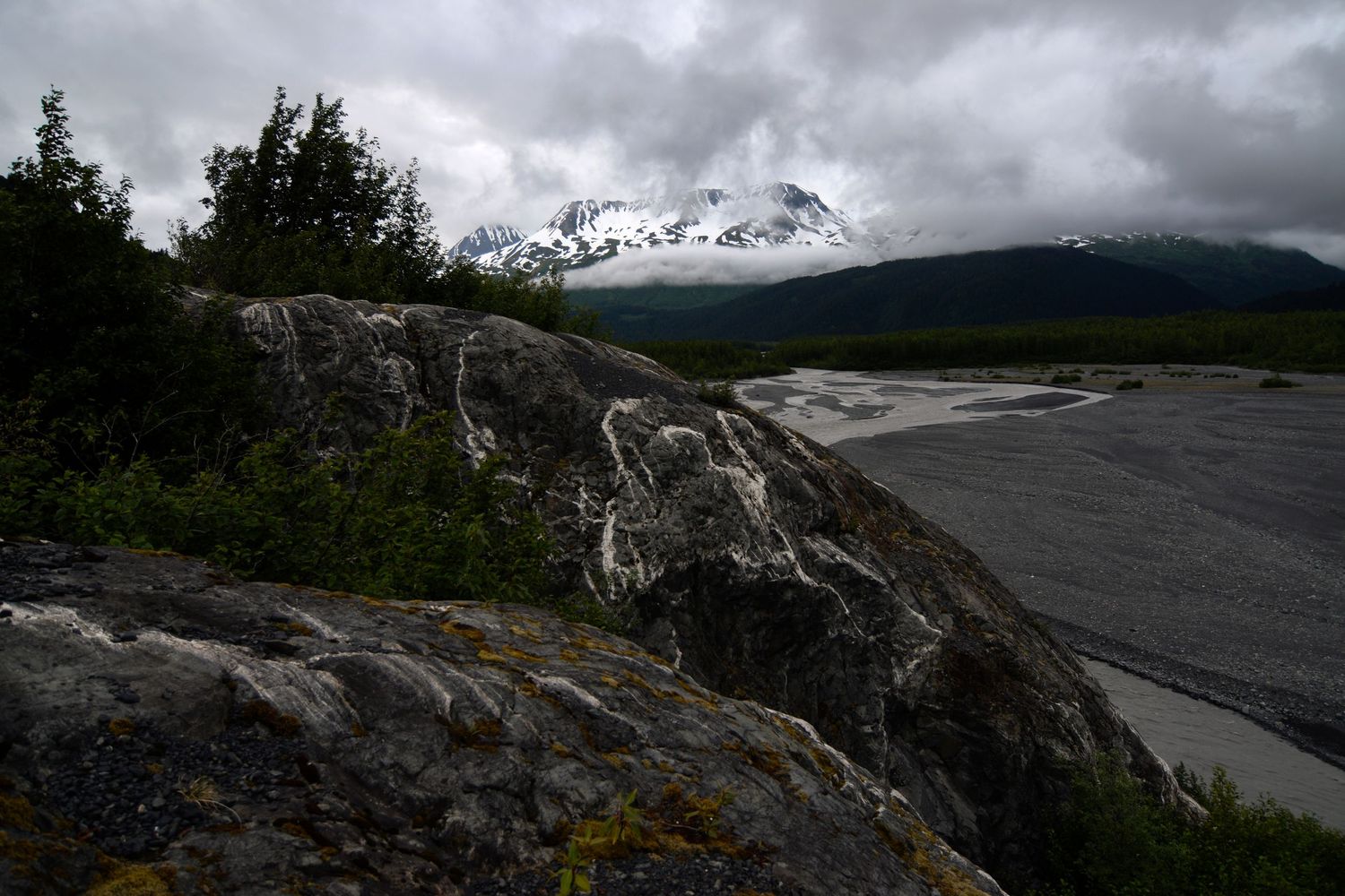 A moody grey overcast day looking at gravel bars past the toe of Exit glacier, with bedrock that has interesting white veins visible.