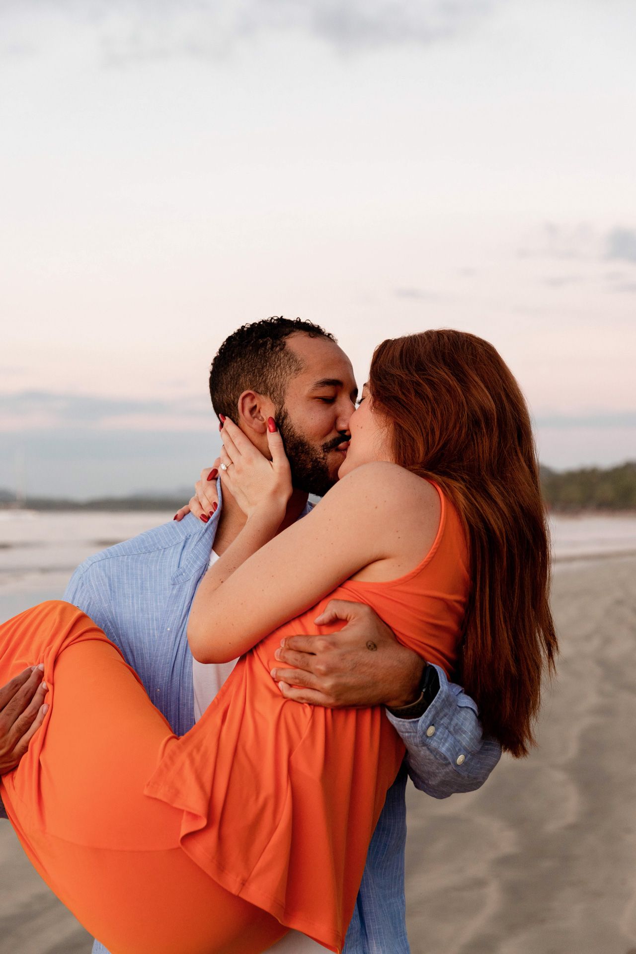 A romantic couple shares a tender kiss on a beach at sunset, with her wearing a flowing orange dress.