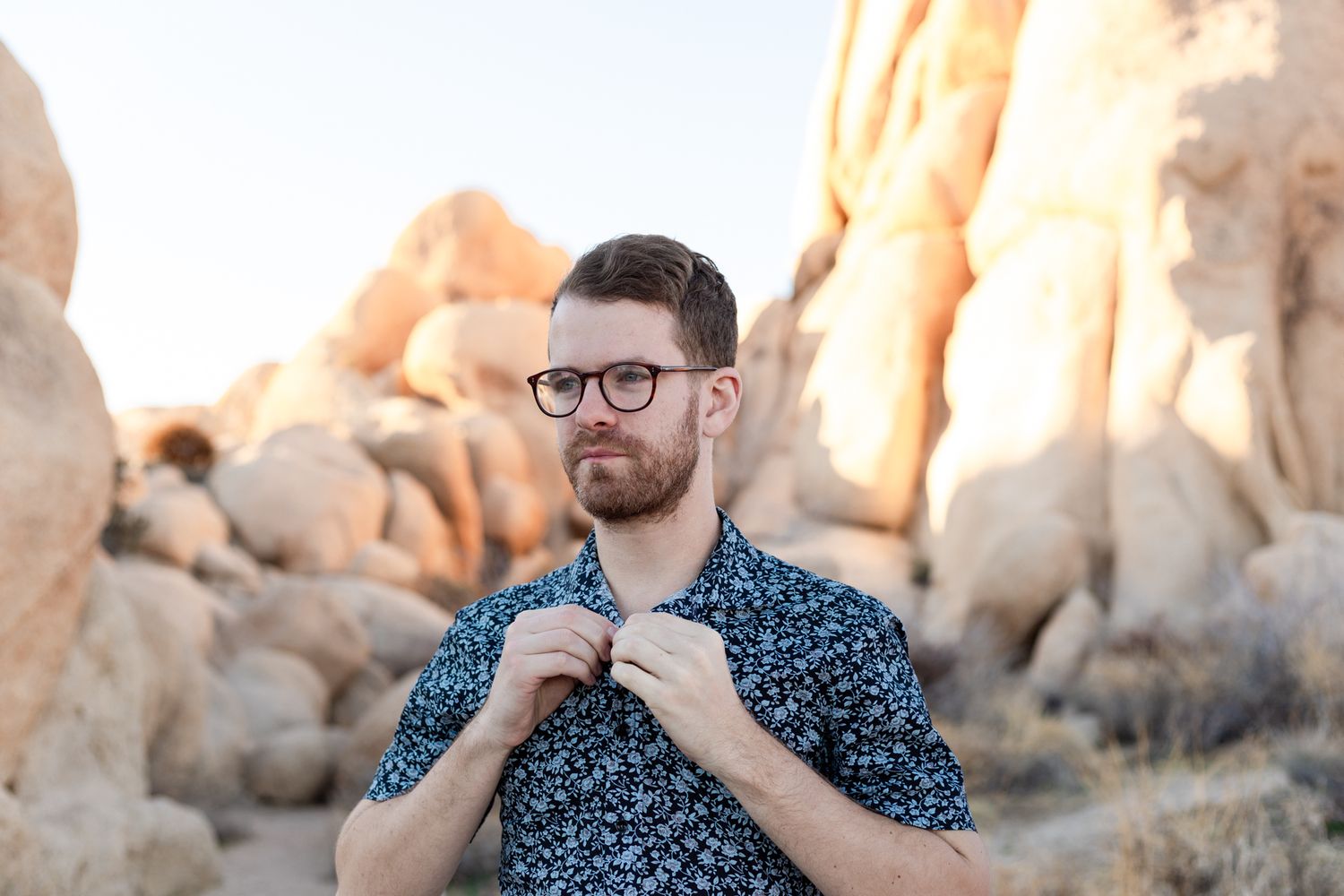 A person wearing a blue polka dot shirt poses in front of desert rock formations at sunset.