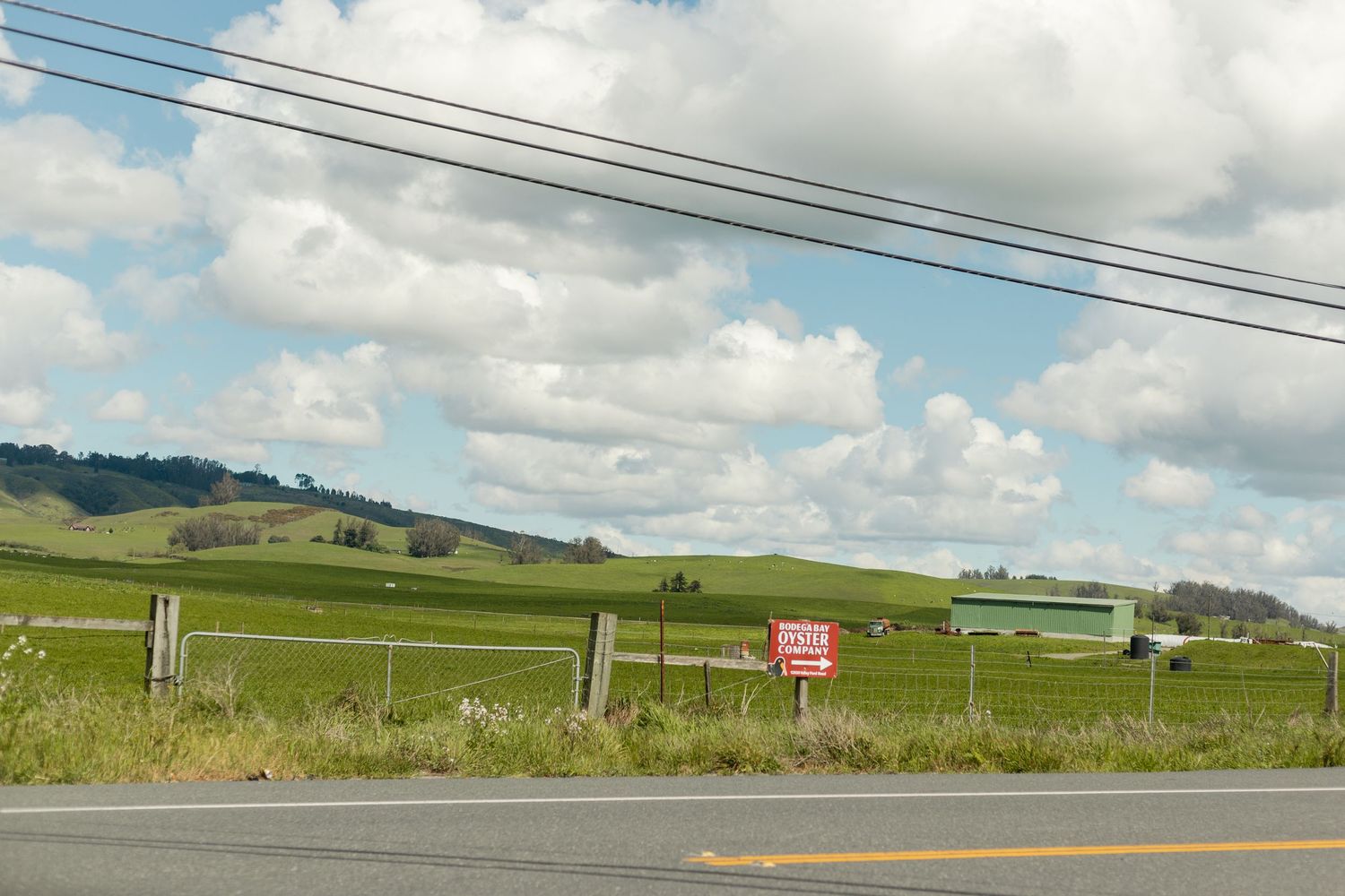Rural countryside road with lush green rolling hills, farmland, a red sign, and power lines under a partly cloudy sky.