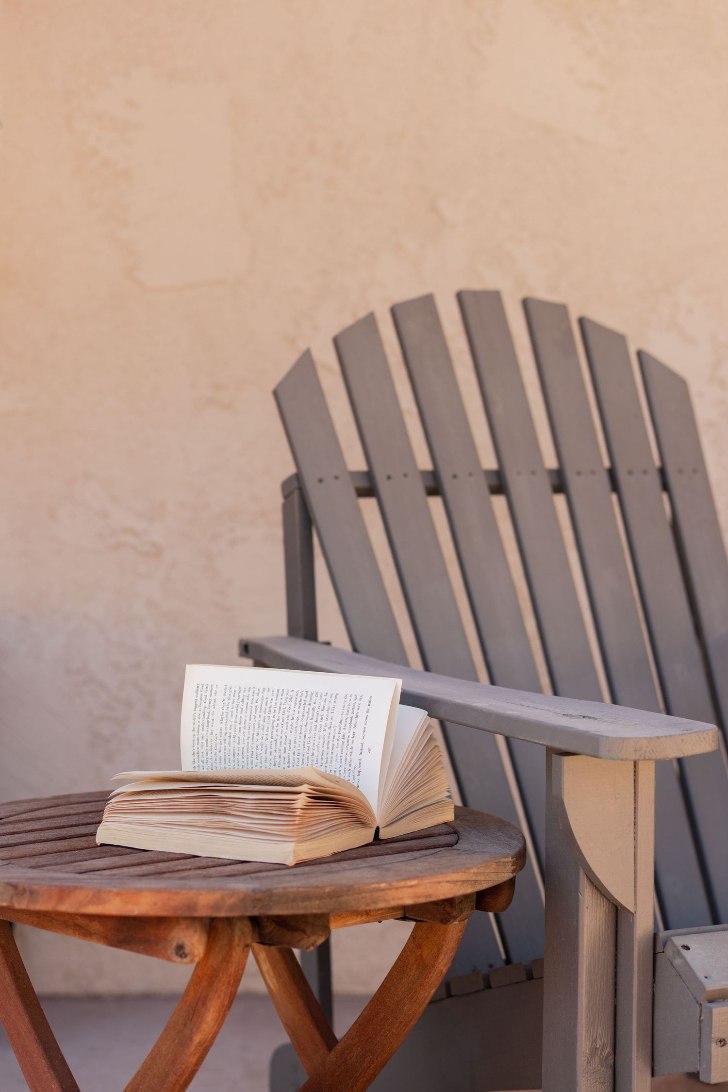 Open book resting on a wooden side table beside a gray Adirondack chair against a beige stucco wall.