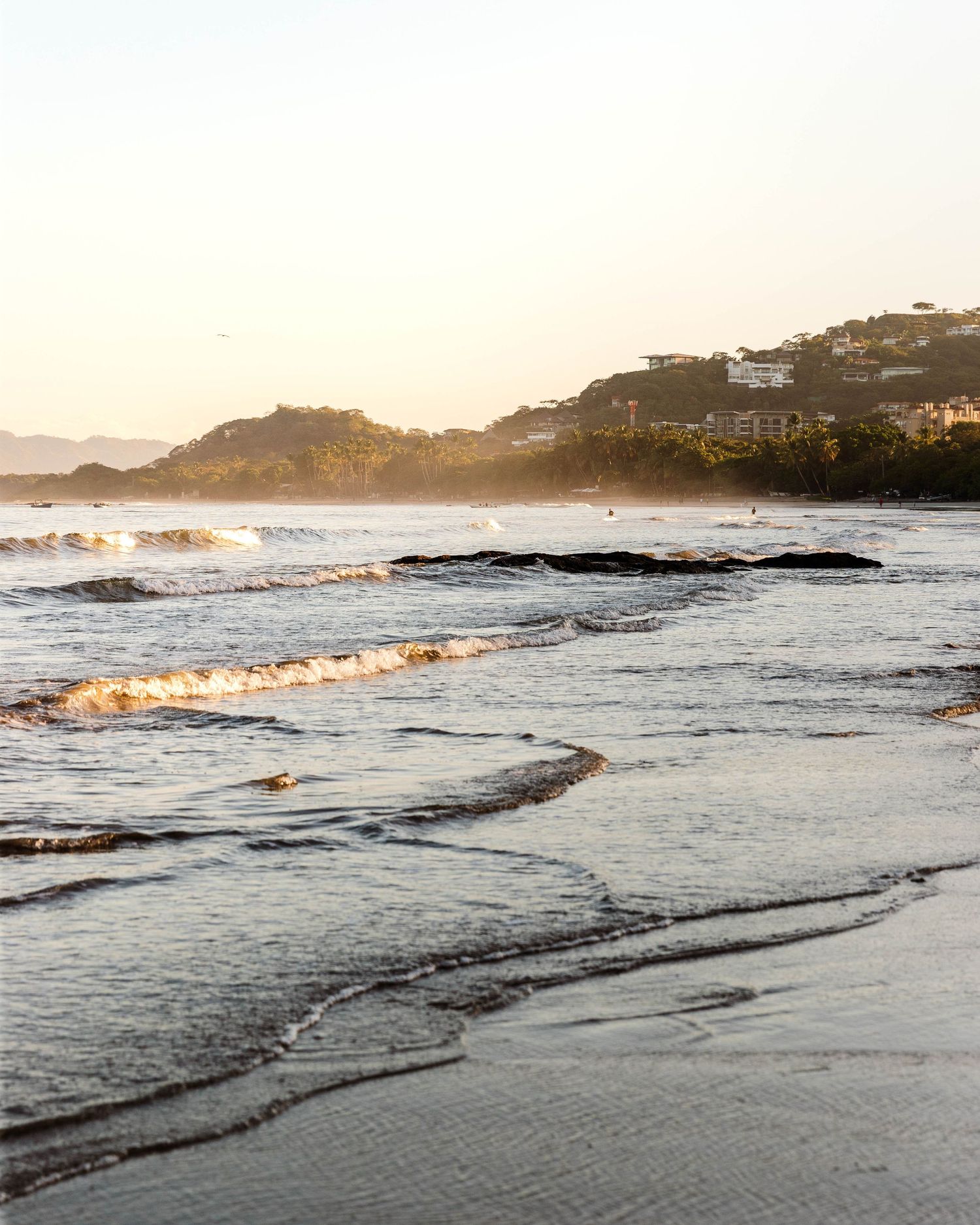 Rocky coastline with waves breaking at sunset against a mountainous backdrop.