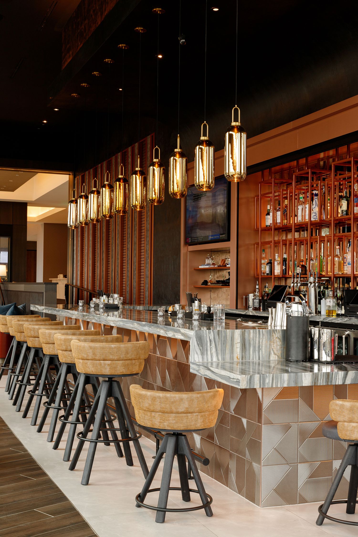 Elegant hotel bar with marble countertop, leather bar stools, gold pendant lights, and backlit shelves stocked with spirits.