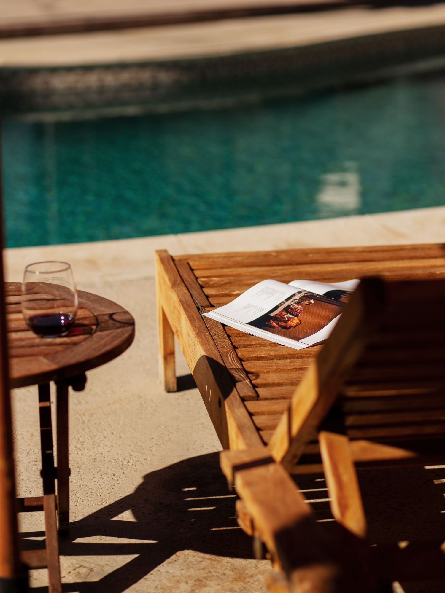 Wooden lounge chair and side table with red wine glass beside a sunny pool, with an open magazine resting on the chair.
