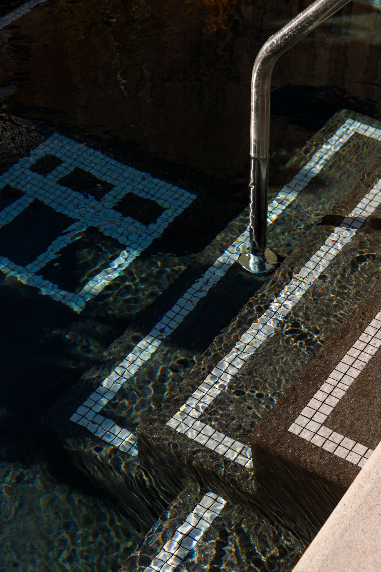 Pool steps with mosaic tile and chrome handrail reflecting sunlight on shimmering blue water.