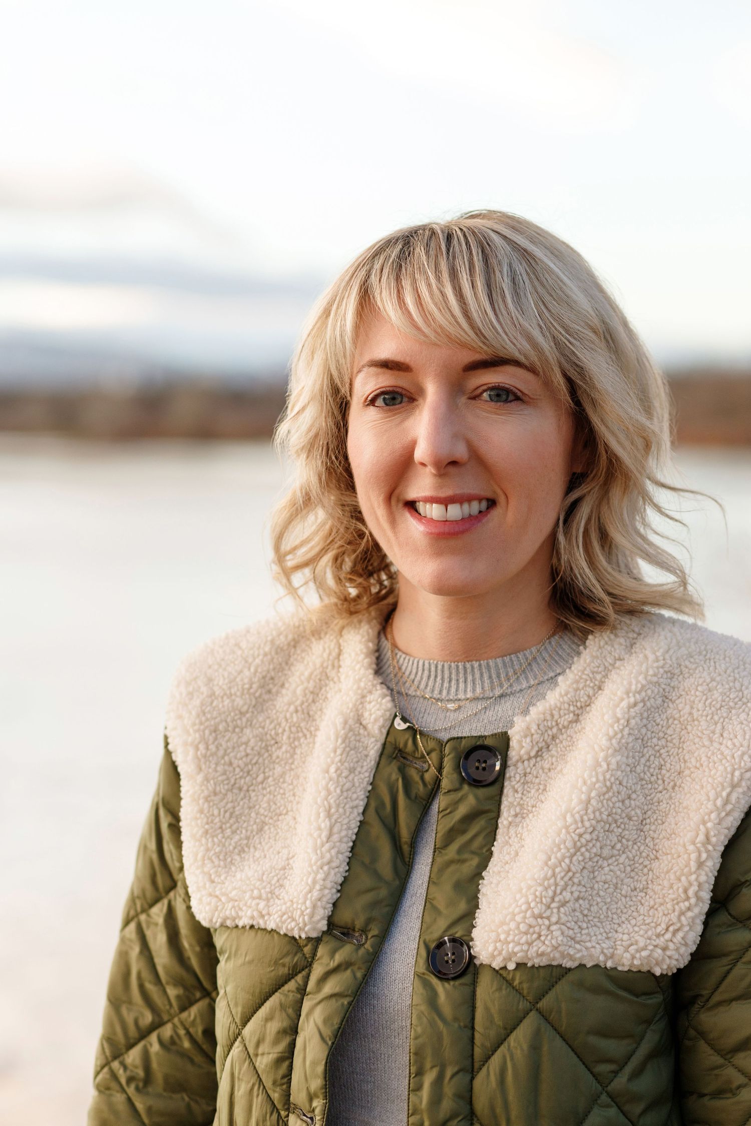 Person in olive quilted jacket with sherpa collar smiles outdoors in winter setting.