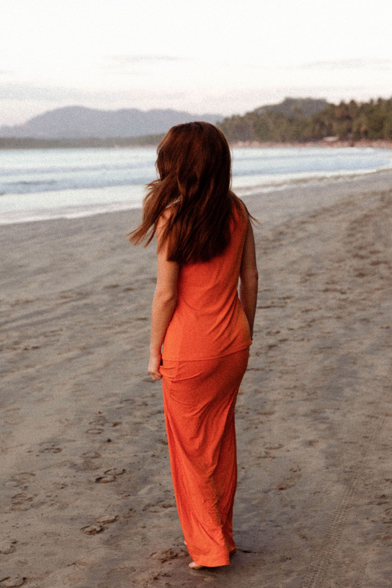Person in flowing orange dress walks along a misty beach shoreline with mountains visible in the distance.