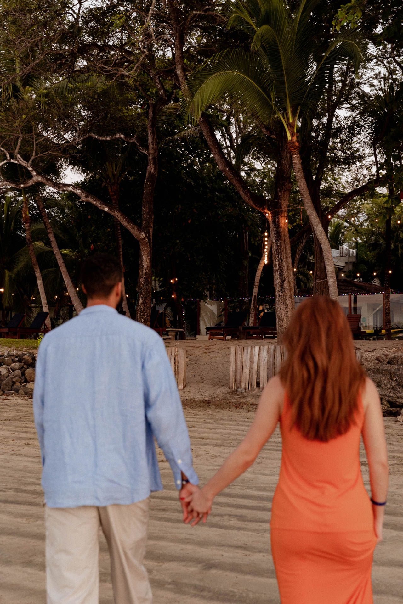 A couple walking hand in hand on a sandy path surrounded by trees at dusk.