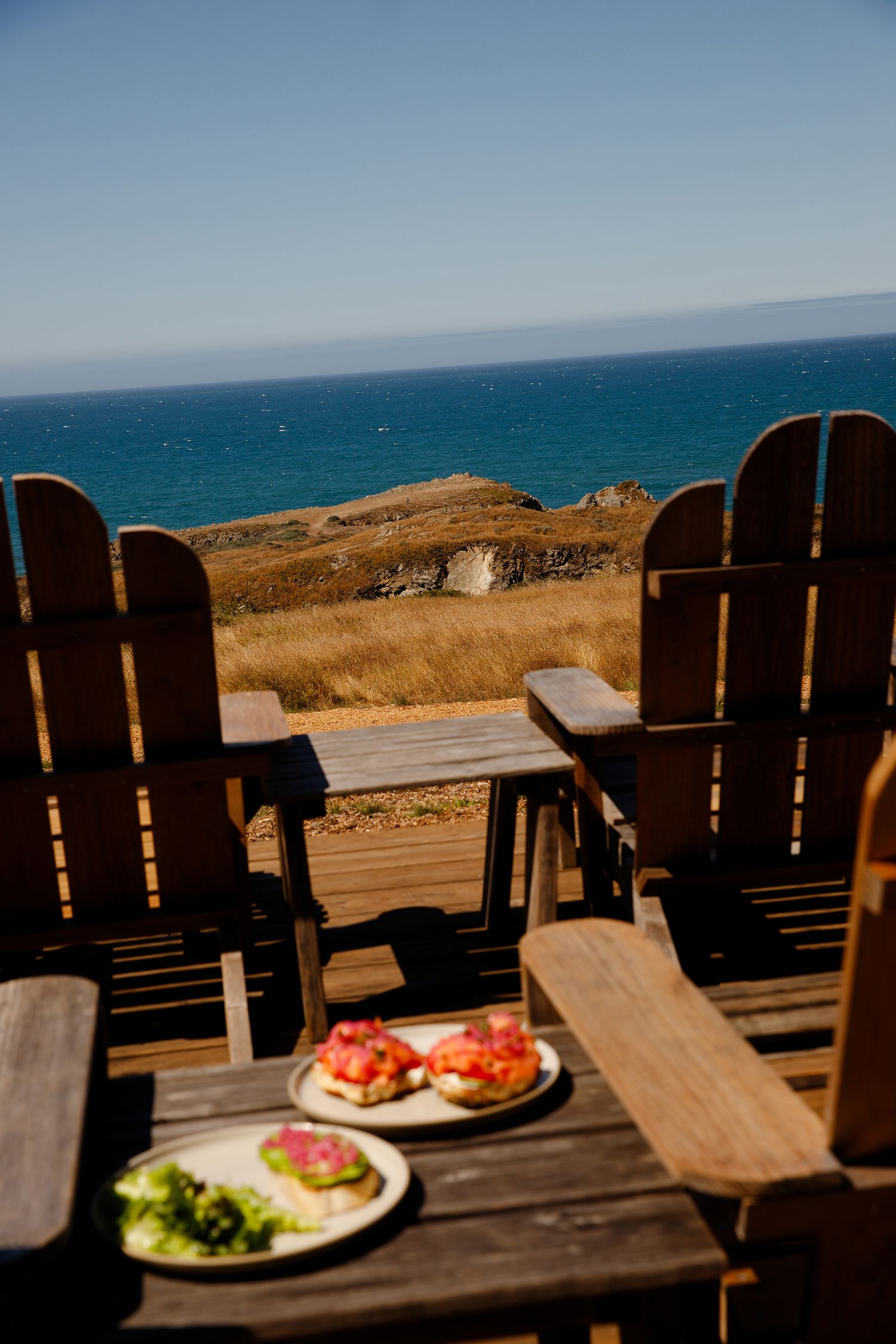 Adirondack chairs with bruschetta plates overlooking coastal cliffs and blue ocean on a sunny day.