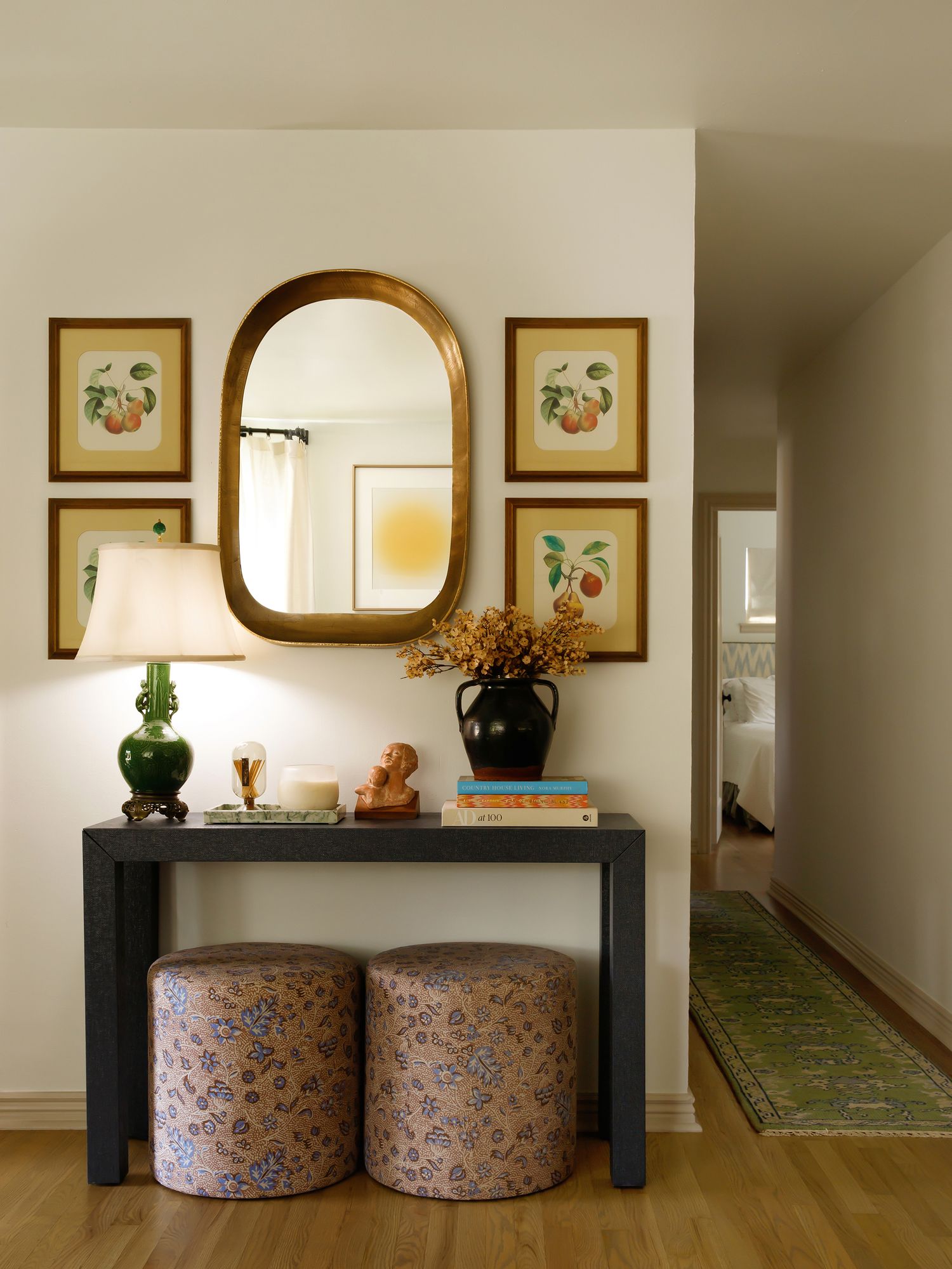 Entryway with black console table, floral ottomans, gold mirror, botanical prints, green lamp, and vase with dried flowers.
