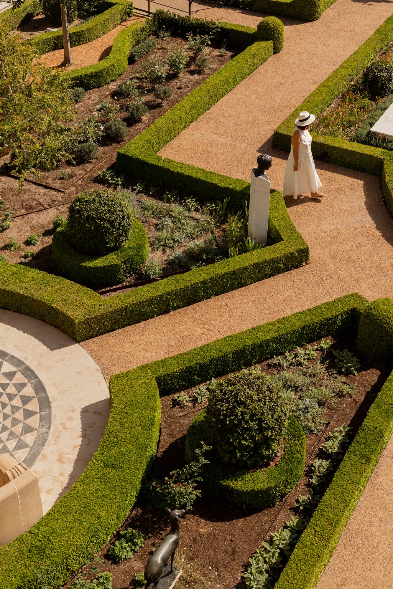 Aerial view of a formal garden with neatly trimmed hedges and a woman in white dress and hat strolling along sandy paths.