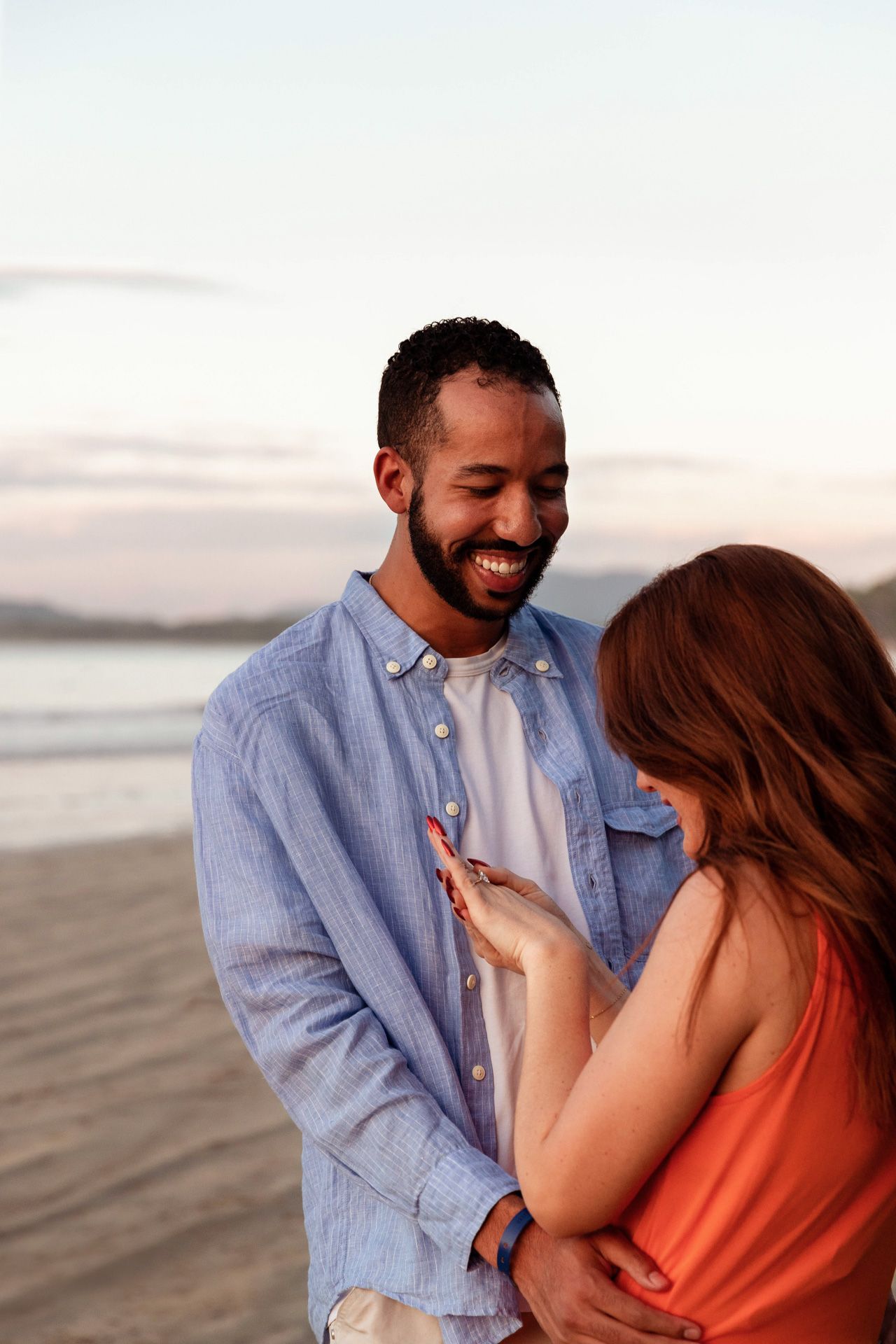 Two people laugh and embrace during a romantic beach photoshoot at dusk.