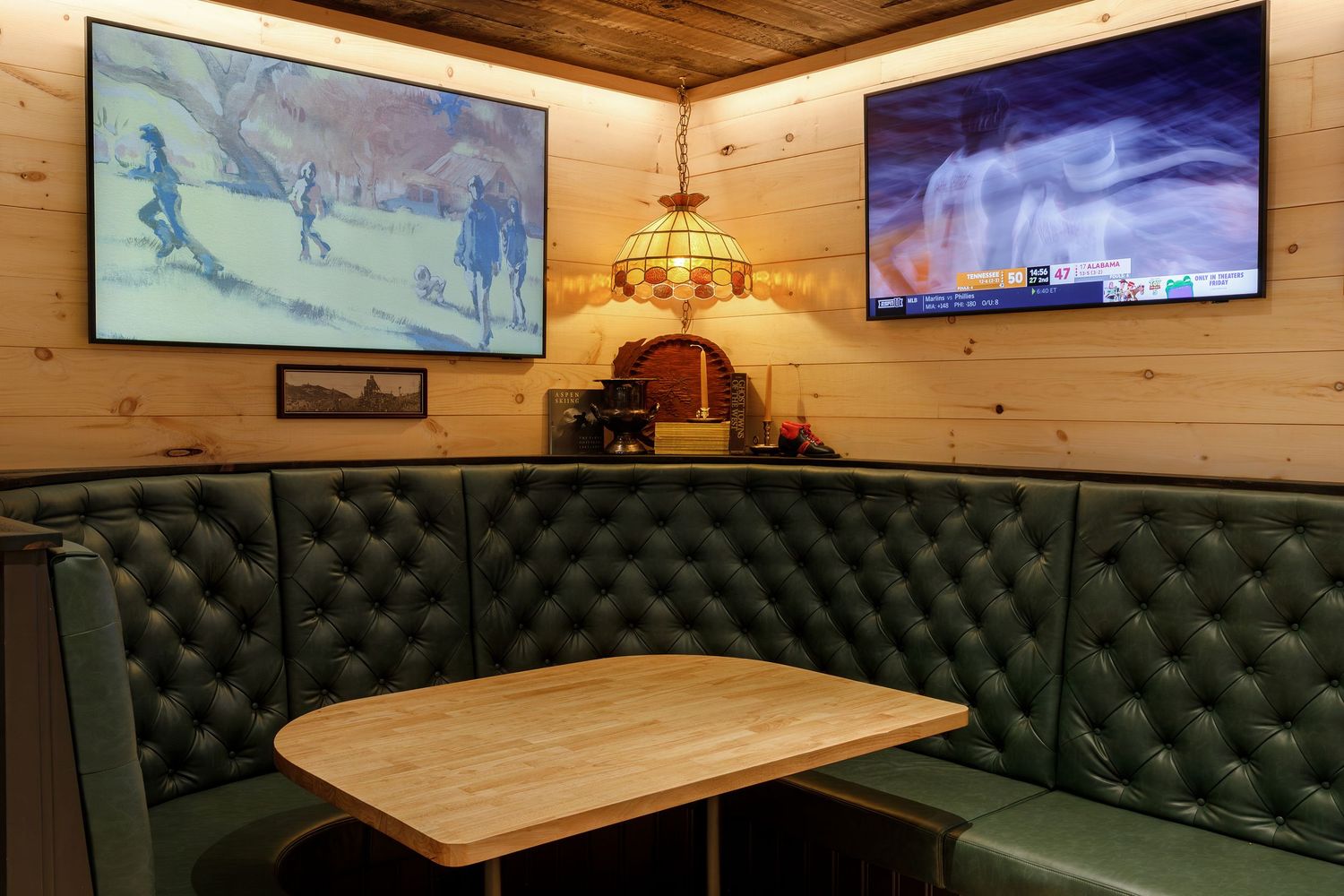 Cozy restaurant booth with green leather seating, wooden walls, two TVs, and a Tiffany-style pendant lamp over a wooden table.