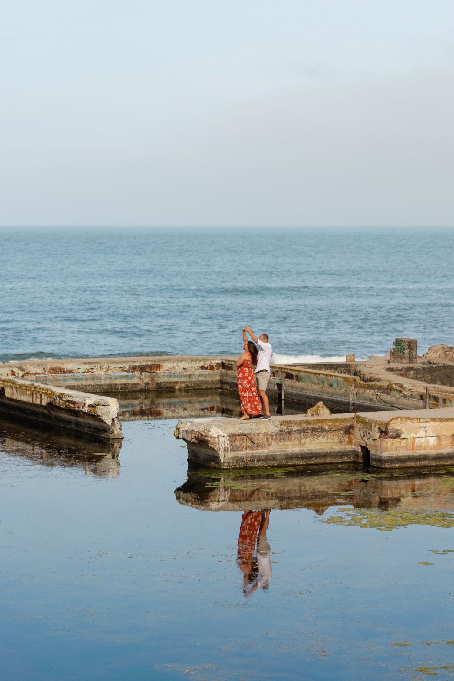 A lone figure stands on a weathered concrete pier extending into the calm blue ocean waters.