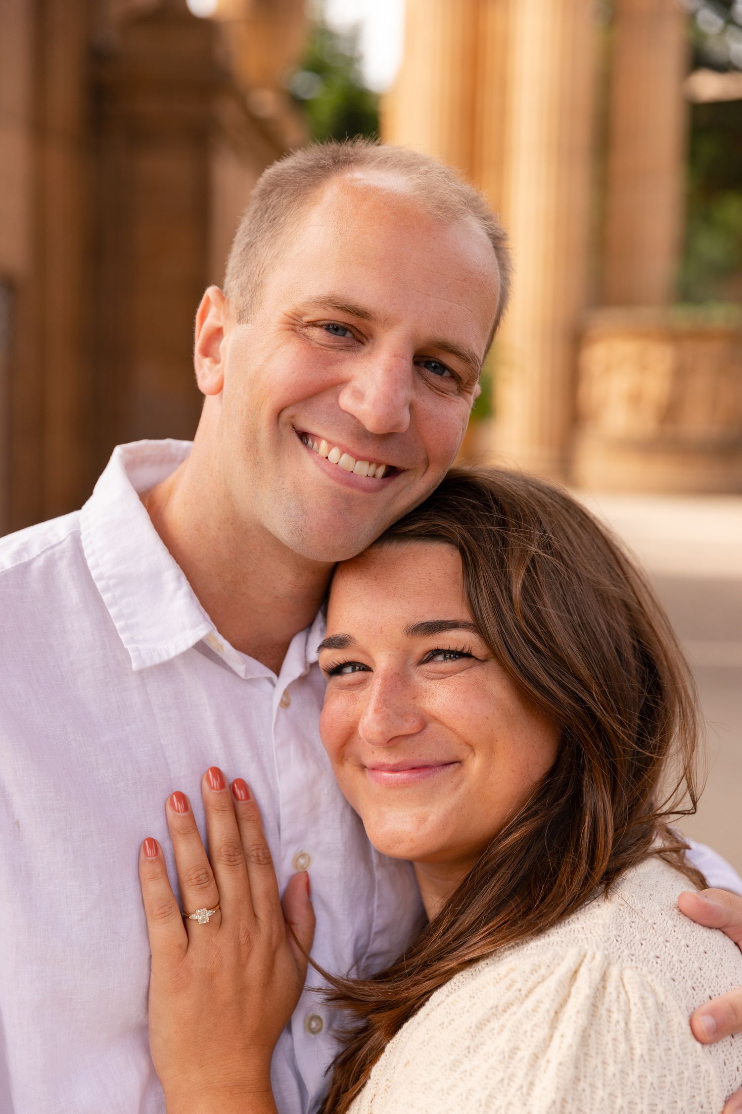 A happy couple embraces and smiles warmly at the camera while standing in front of outdoor pillars.
