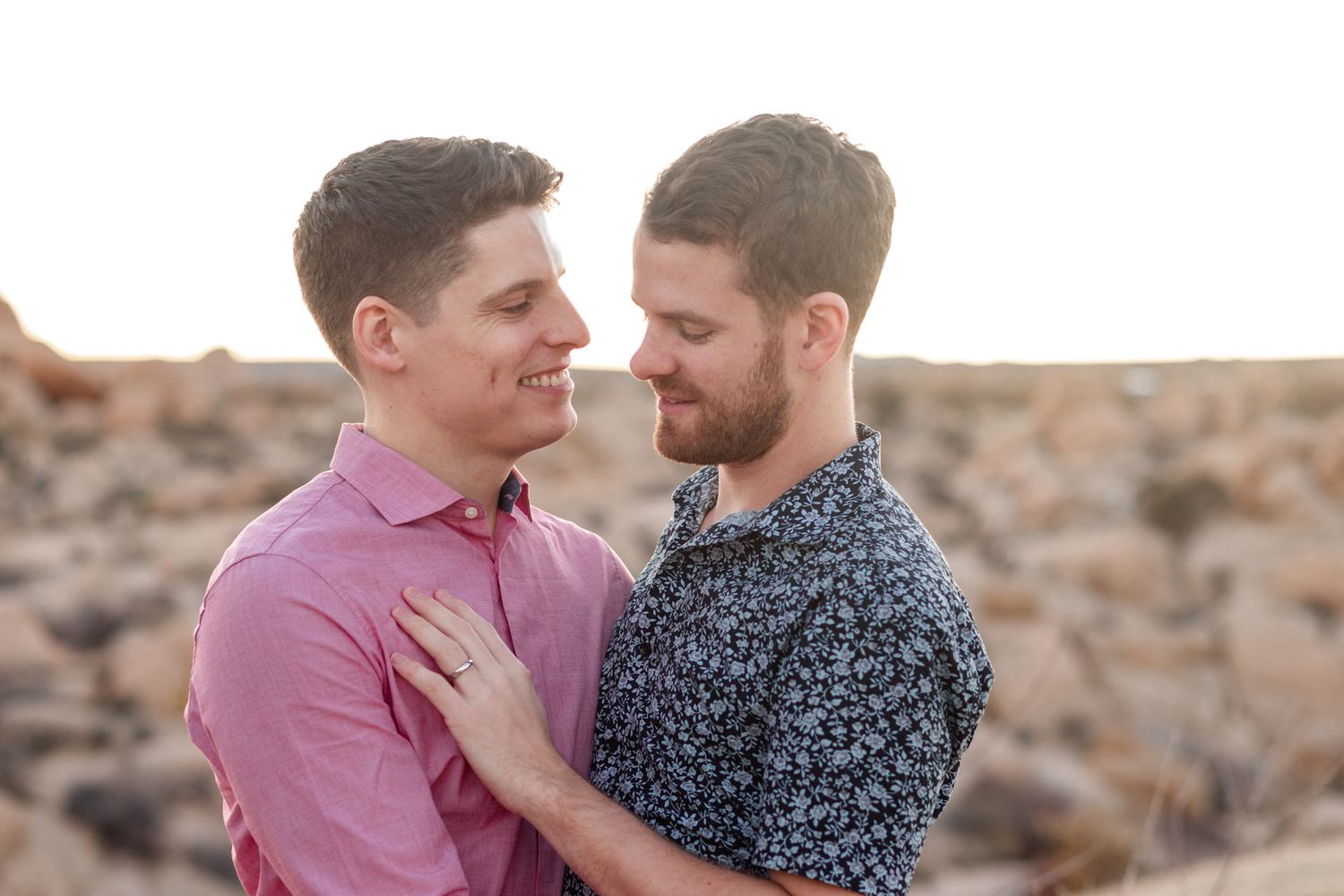 A couple in pink and navy patterned shirts embrace lovingly in front of a stone wall.