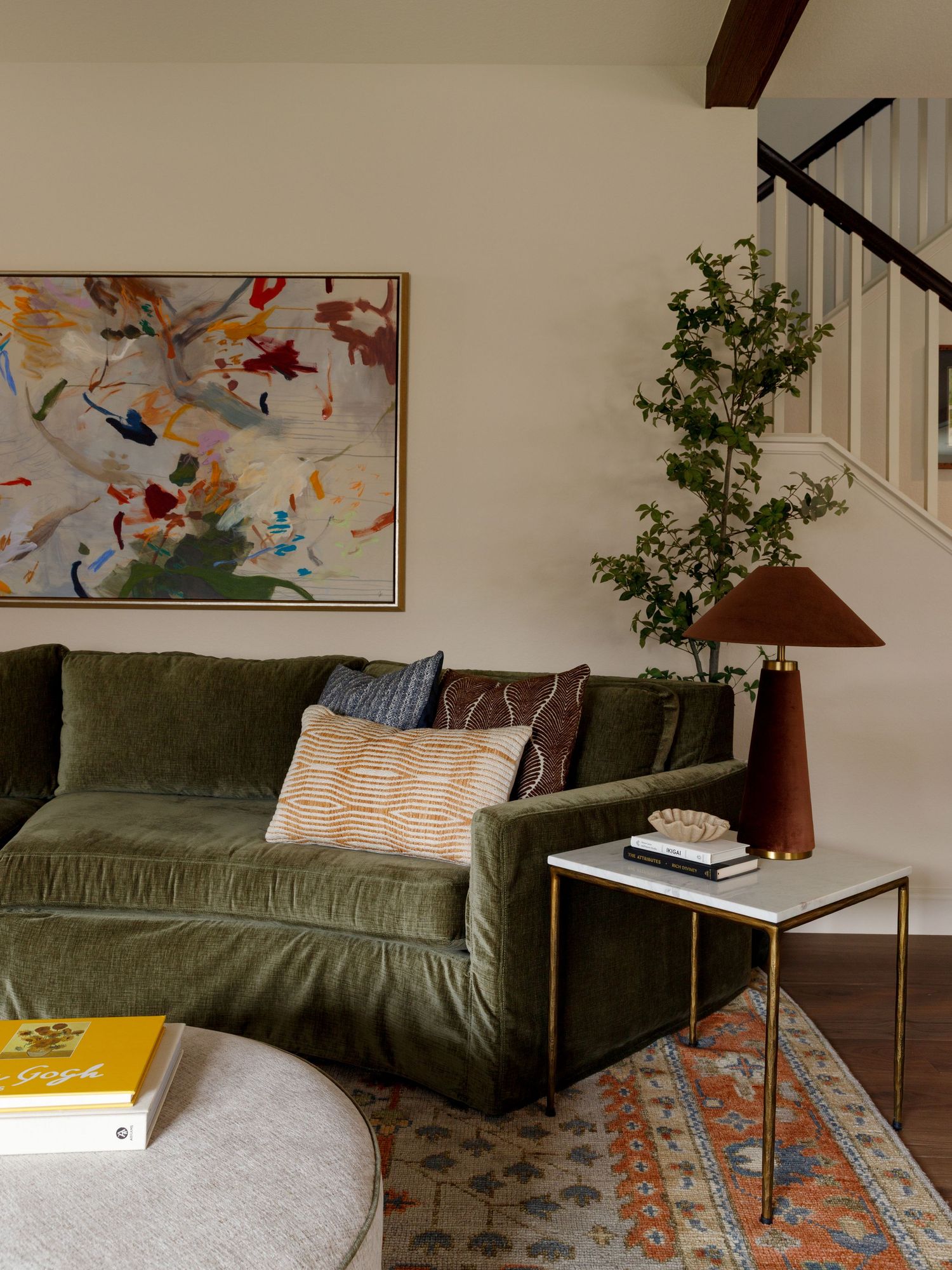 Green velvet sofa with colorful abstract art, terracotta lamp, patterned rug, and indoor tree in a cozy living room.