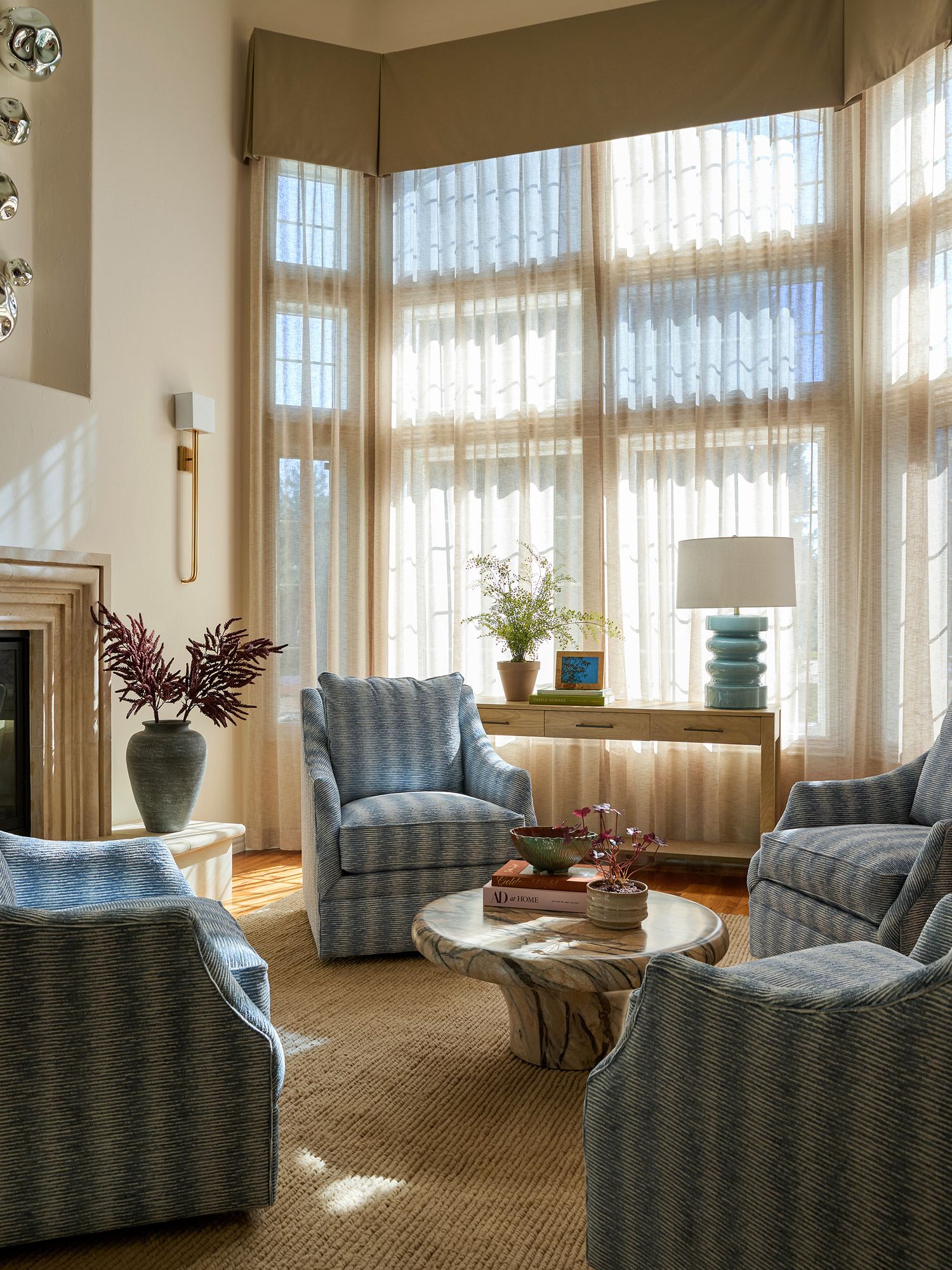 Elegant living room with blue striped armchairs, round wood coffee table, bay windows with sheer curtains, and a teal lamp.