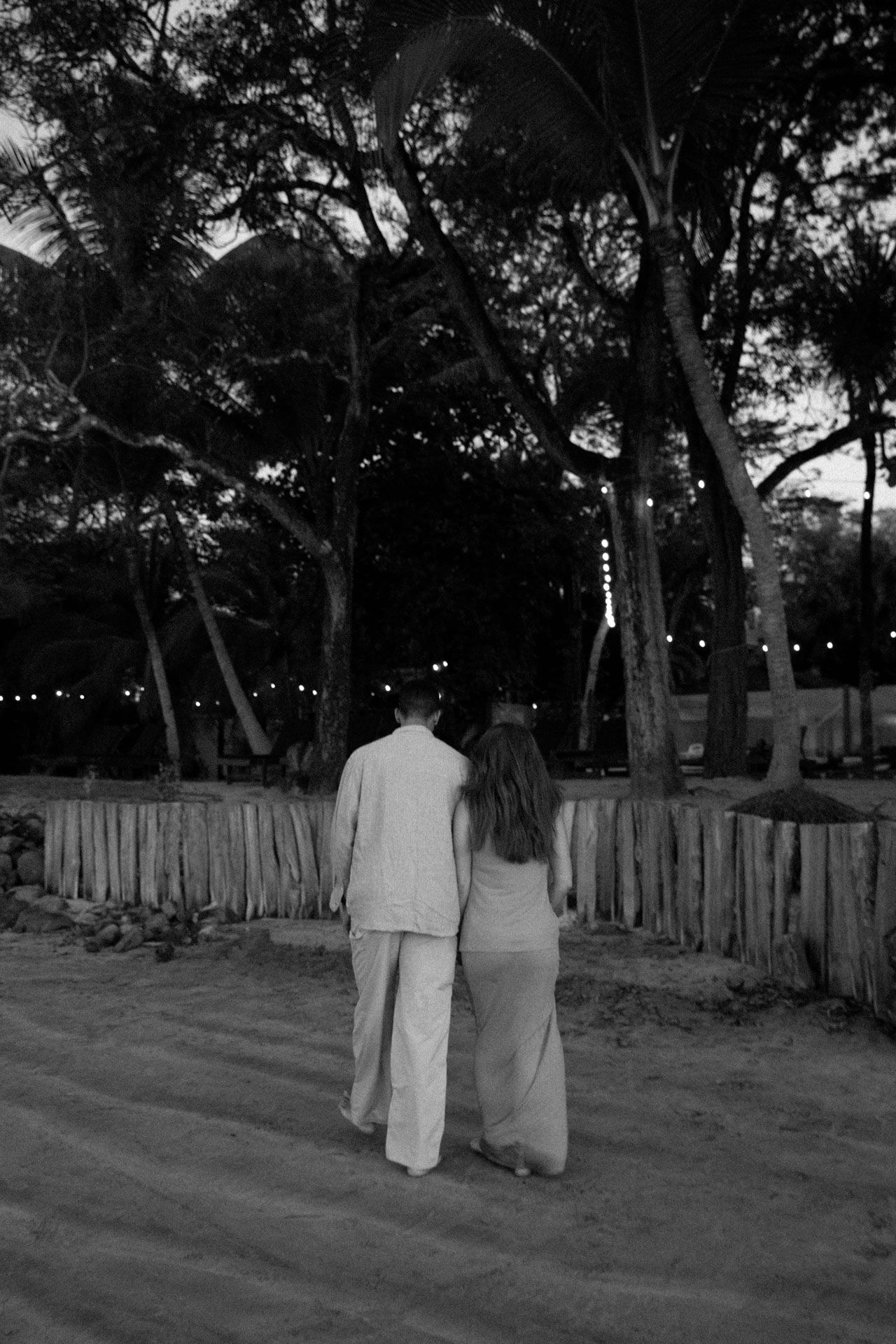 Two people walk together along a path at dusk with a wooden fence and trees silhouetted against the darkening sky.