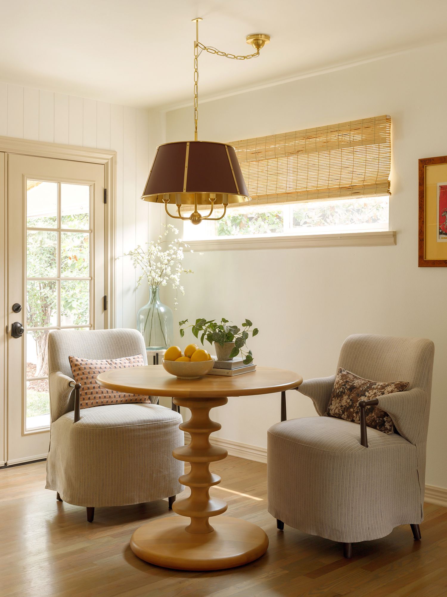 Cozy breakfast nook with round wood pedestal table, gray slipcovered chairs, brass pendant light, and bamboo roman shade.
