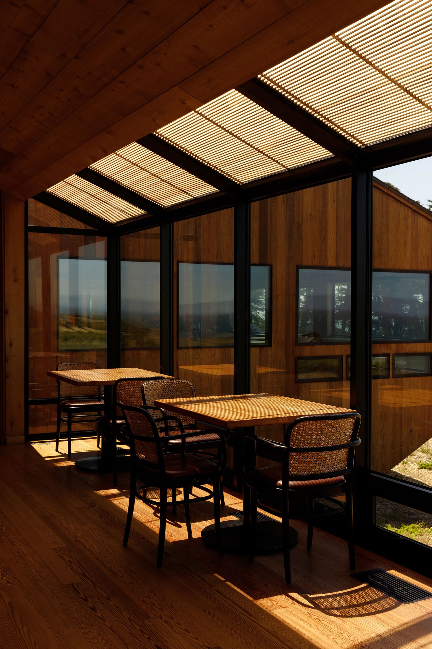 Sunlit wooden dining area with tables and chairs in a glass-walled room with slatted ceiling.
