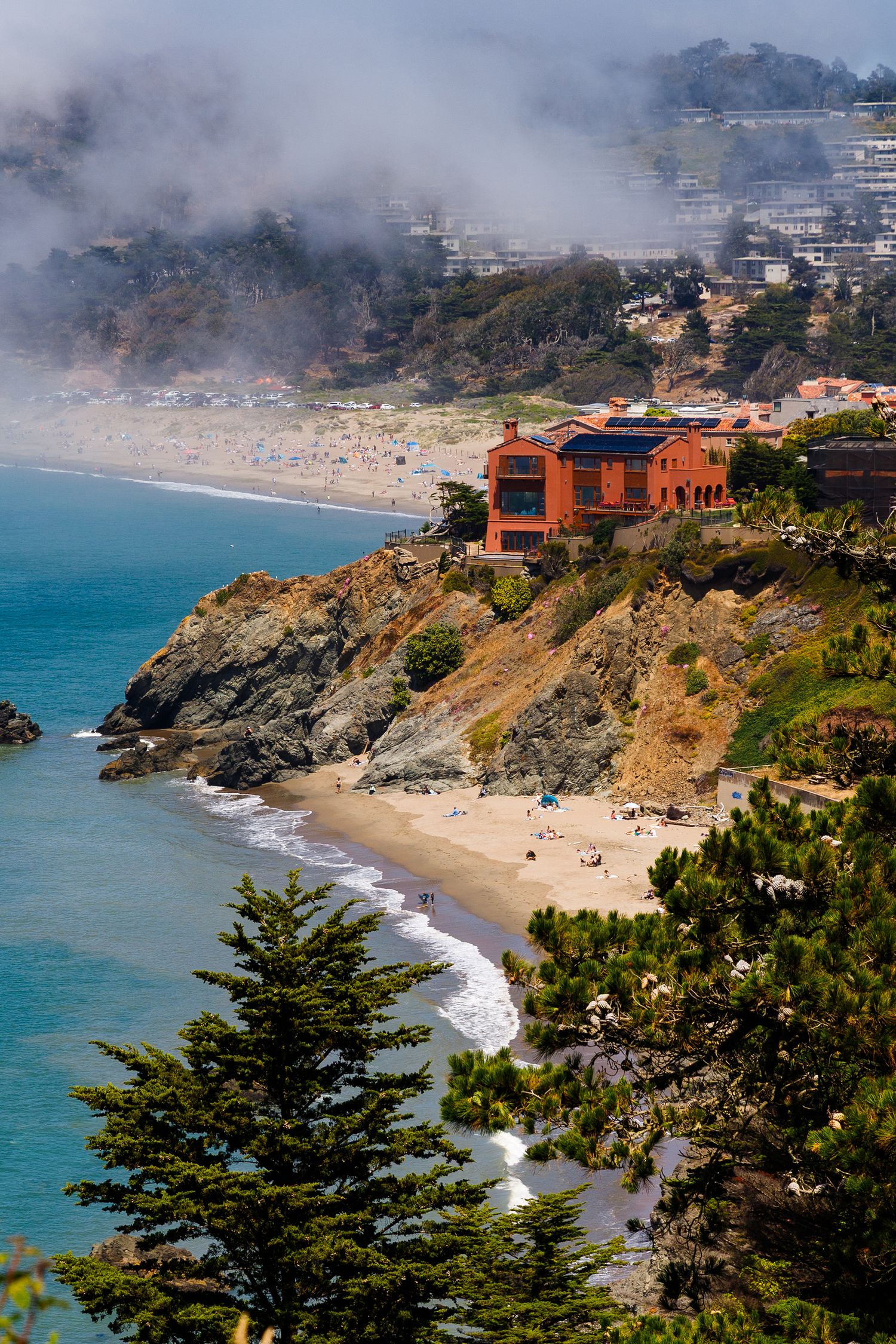 Aerial view of a red building on rocky cliffs overlooking a sandy beach with fog rolling in over the California coastline.