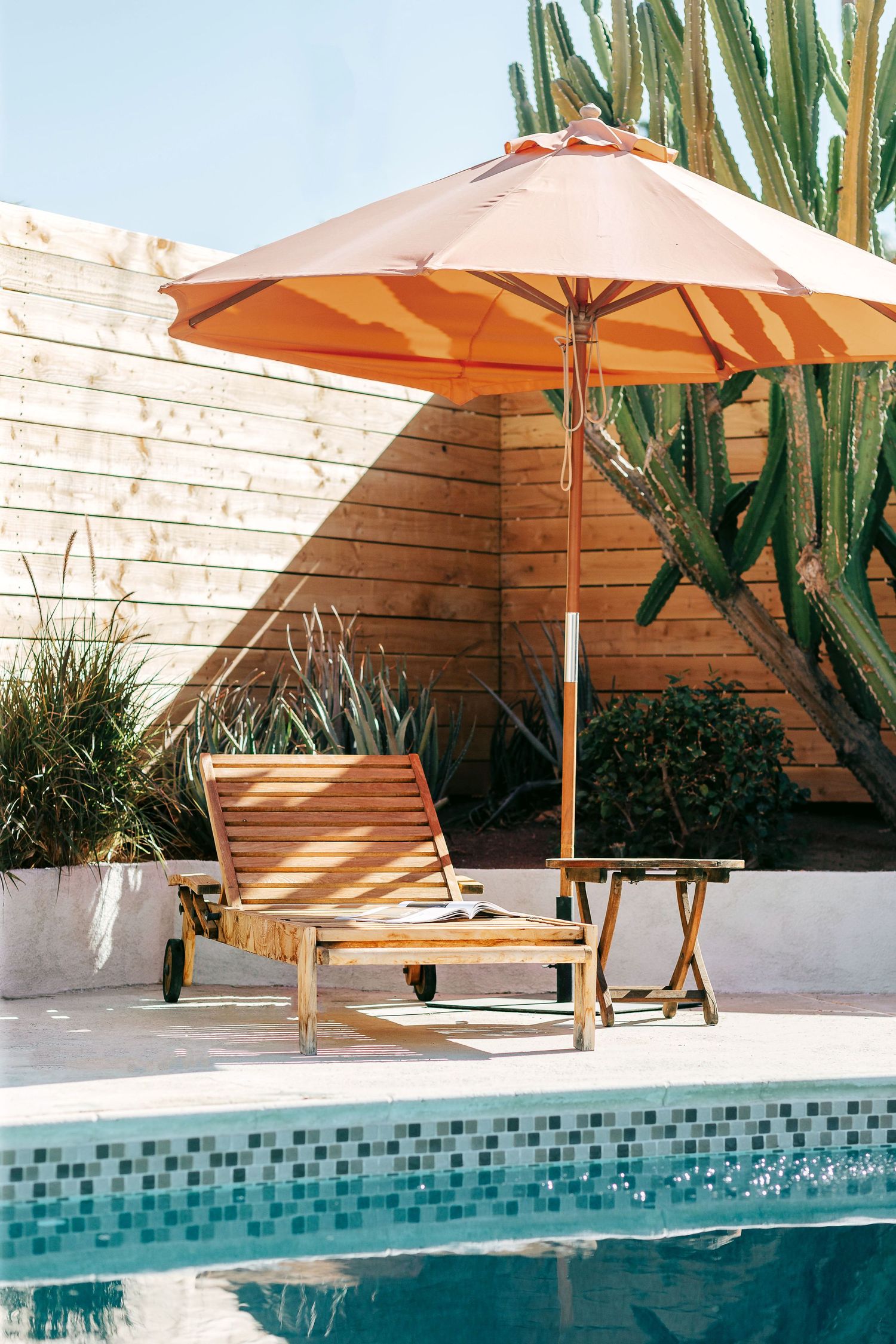 Poolside lounge chair in Palm Springs vacation home with orange umbrella next to tall cacti and wooden fence shot by interior photographer