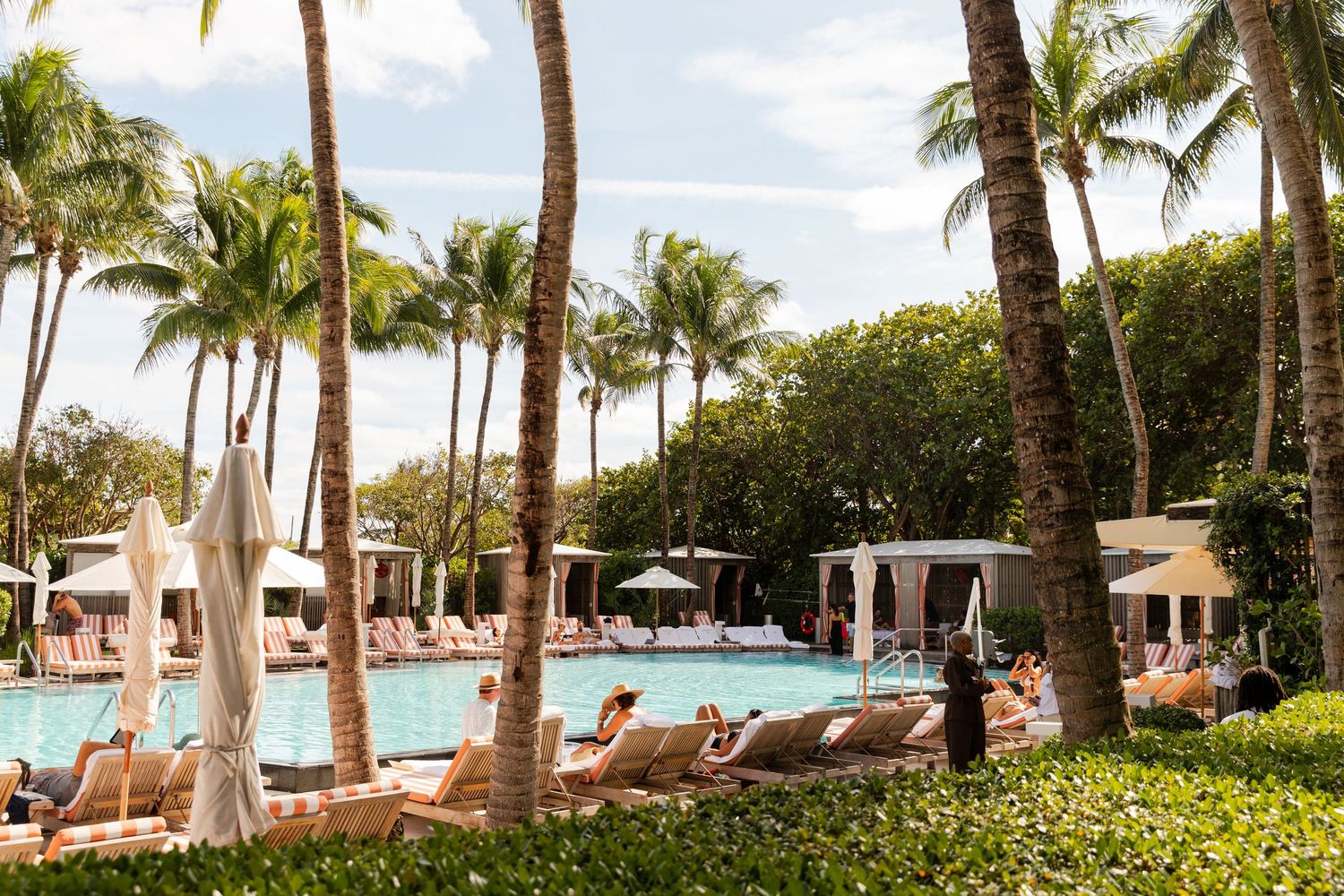 Luxury resort swimming pool surrounded by tall palm trees and white cabanas with lounge chairs on a sunny day.
