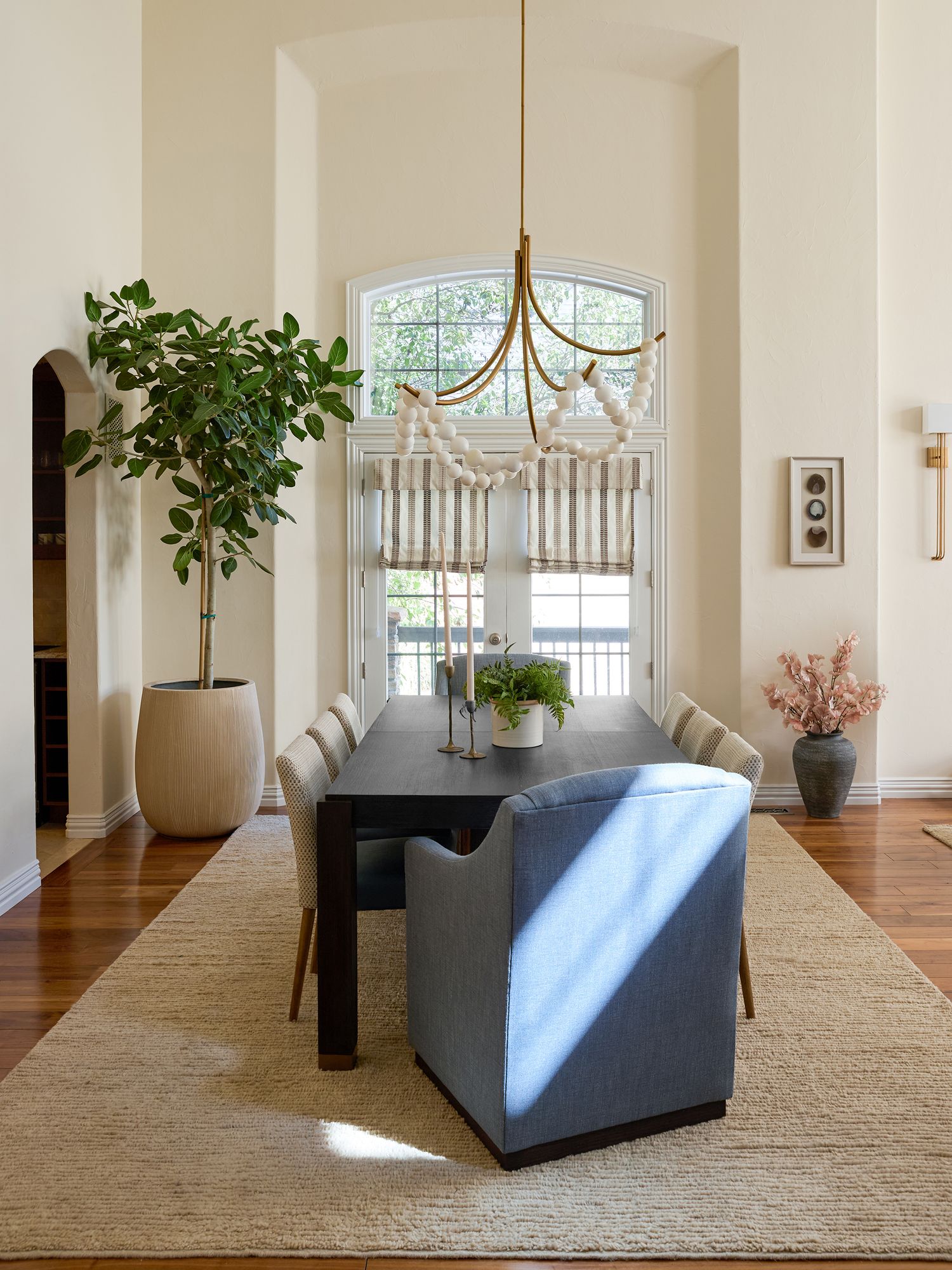Elegant dining room with dark wood table, blue bench, gold chandelier, arched window, and fiddle leaf fig tree.