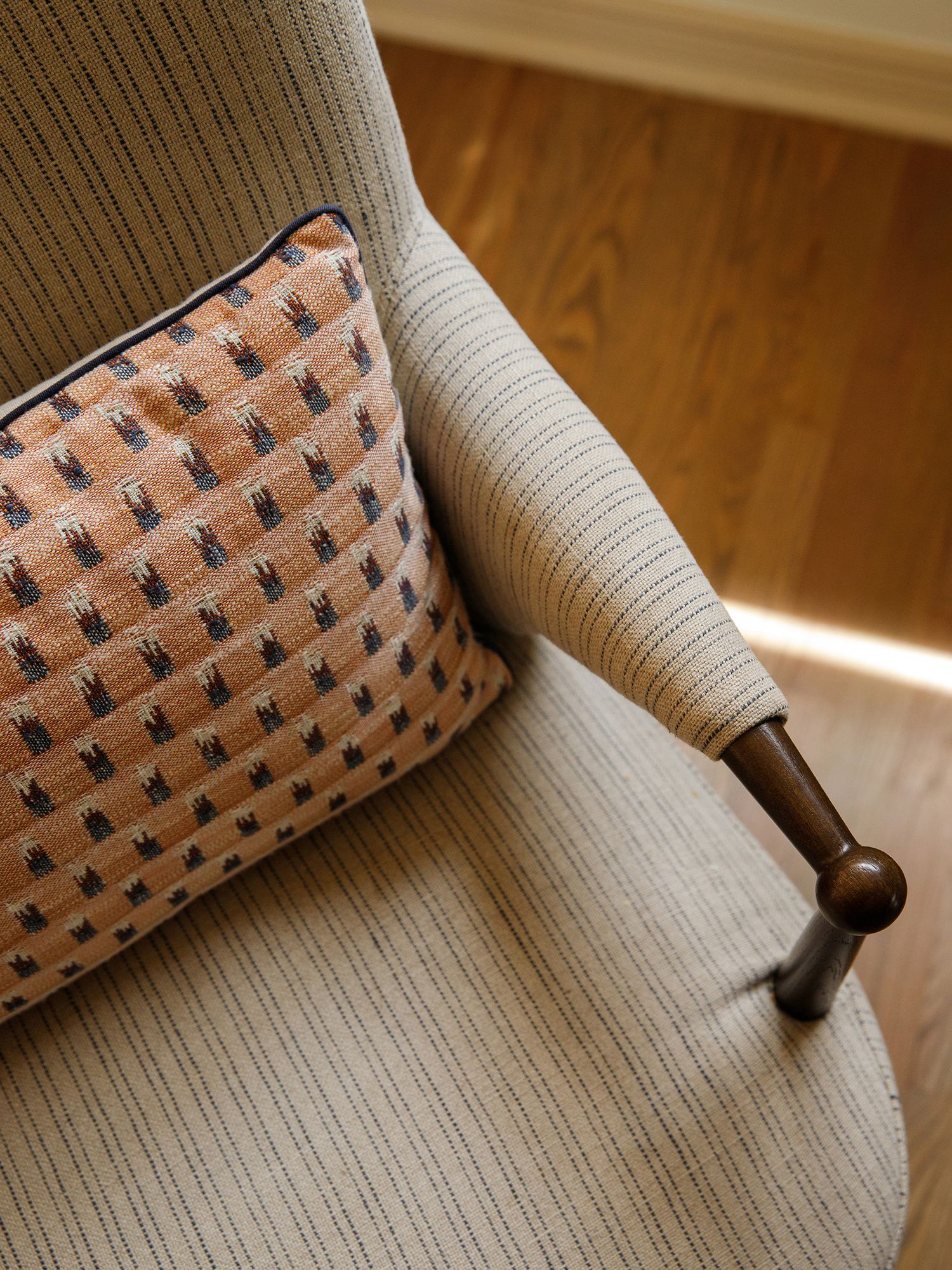 Close-up of a beige striped armchair with a wooden arm detail and an orange patterned throw pillow on hardwood floors.