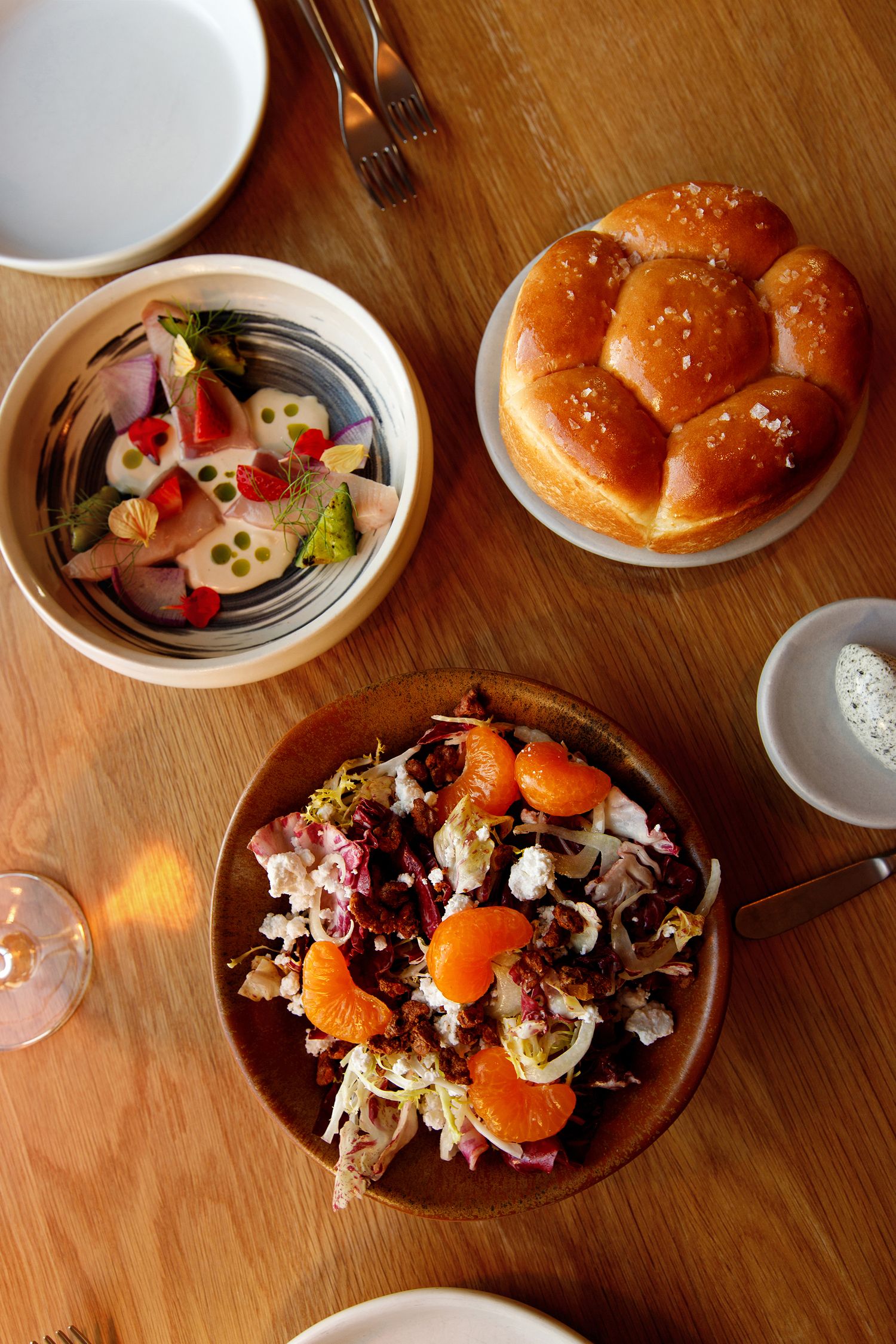 Overhead view of restaurant table with brioche bun, citrus salad, and colorful appetizer bowl on wood surface.