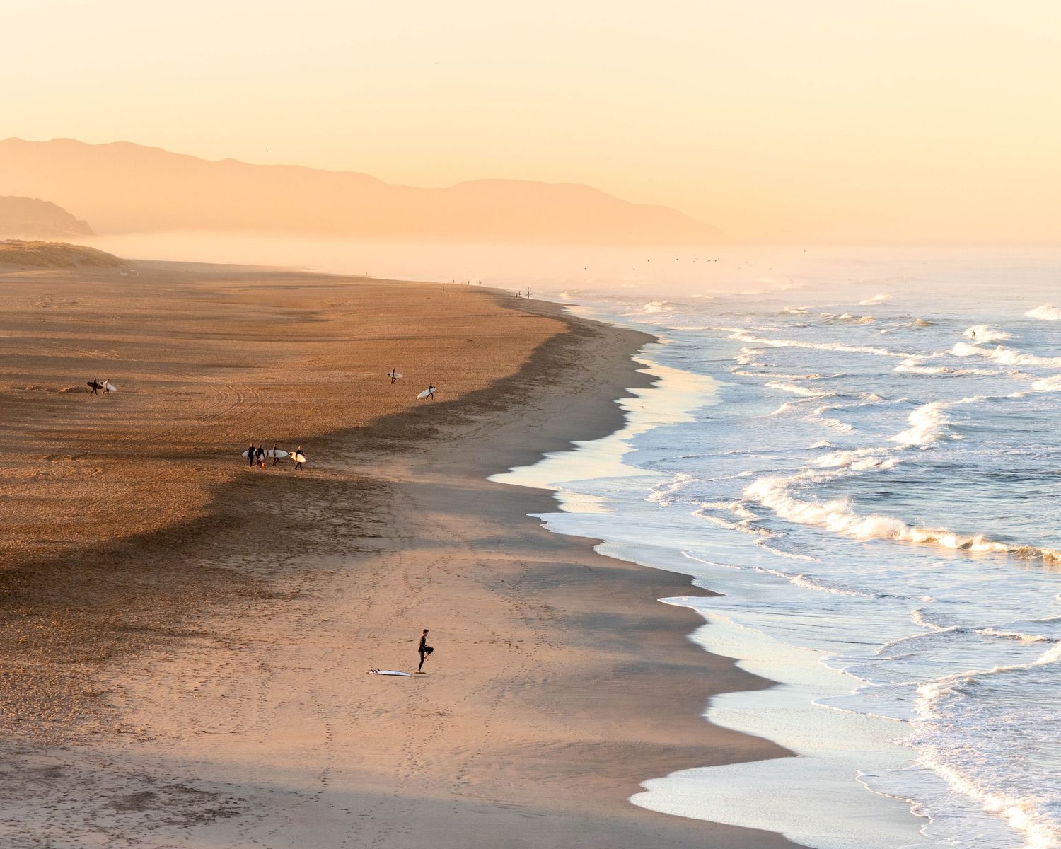 Editorial travel photography image of lone figure walking on misty beach at sunrise with golden sand and rolling waves.