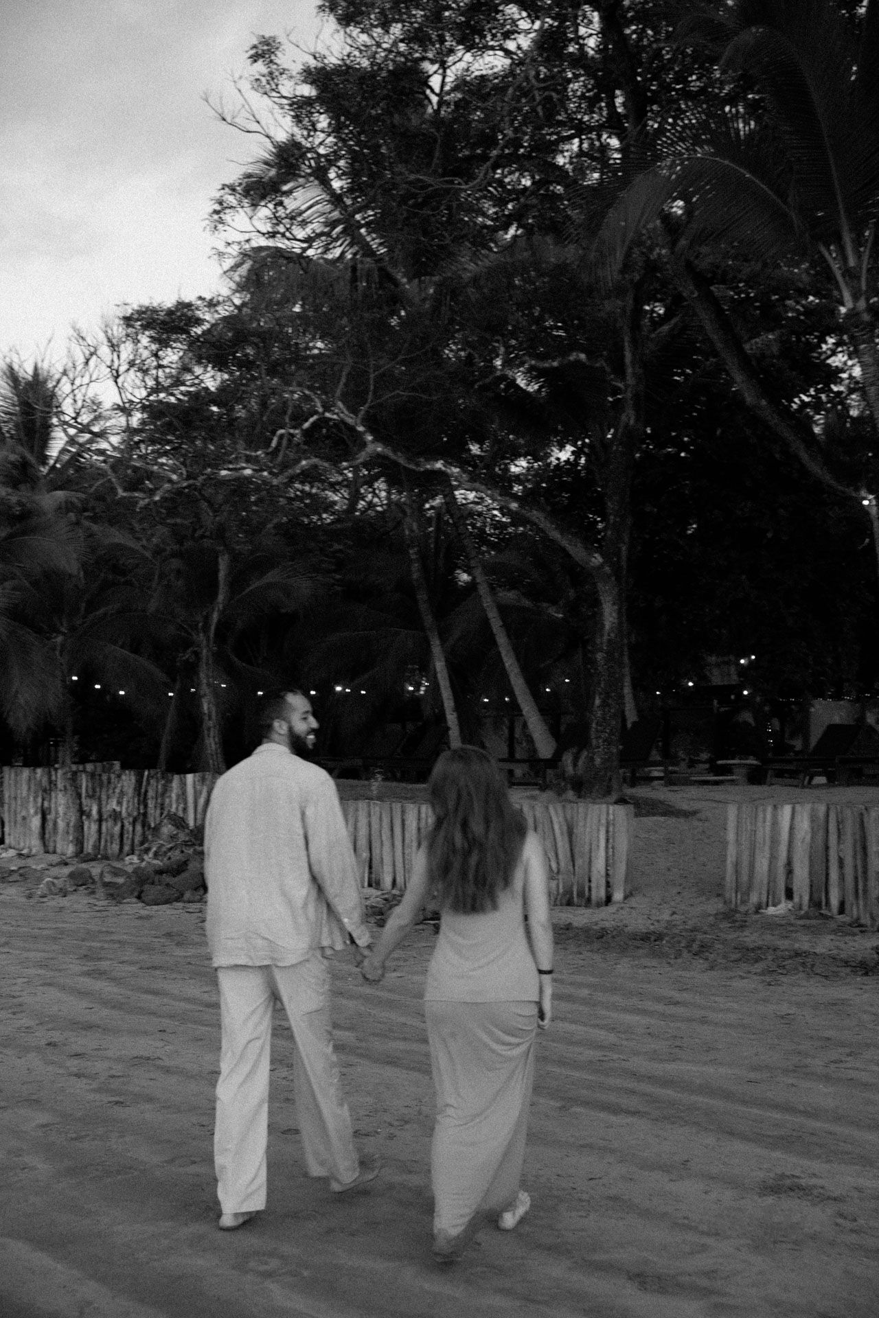 A couple takes a romantic evening stroll along a beach path with trees and string lights in the background.