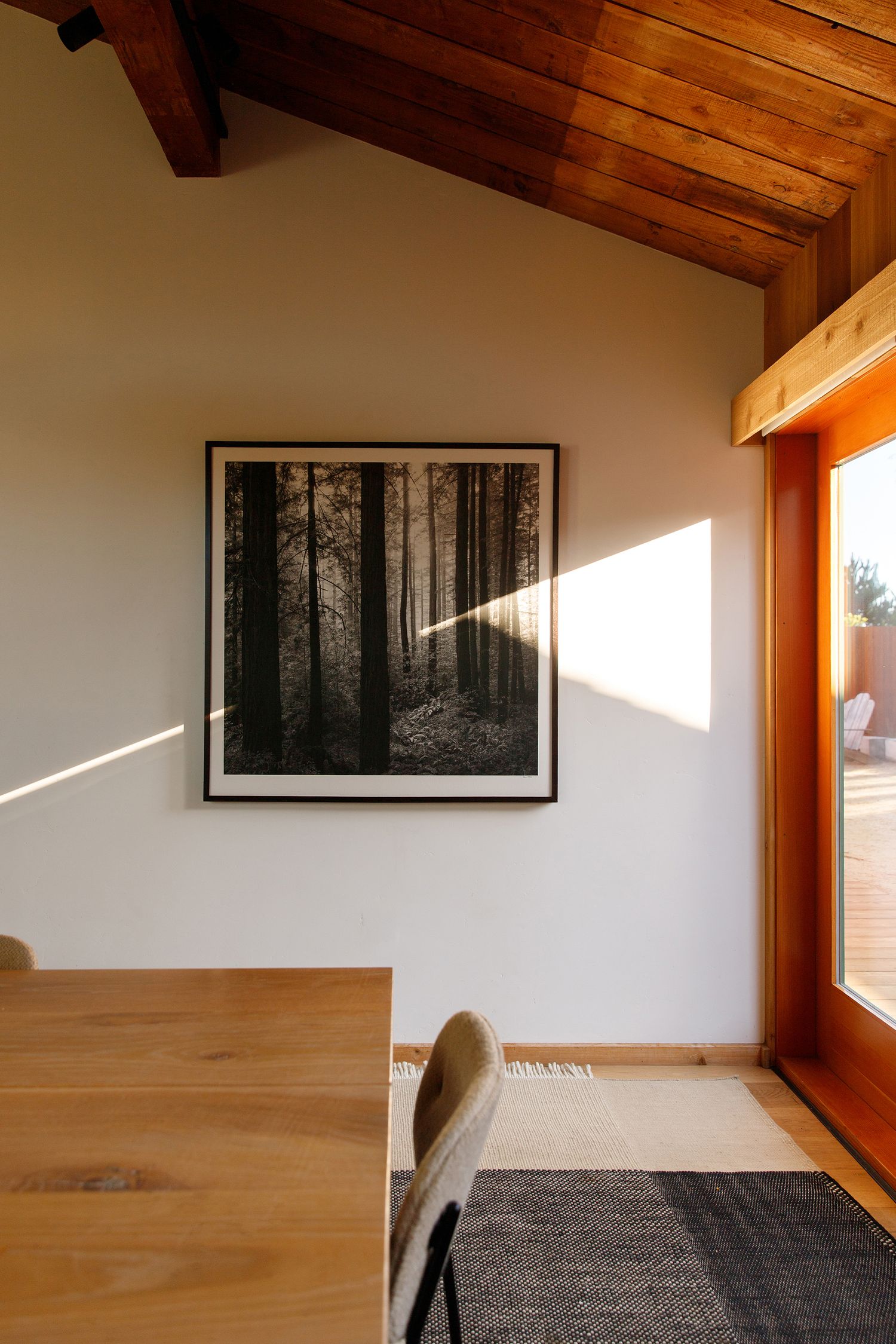 Minimalist dining area with wood ceiling, framed black-and-white forest print on white wall, natural light from glass door.