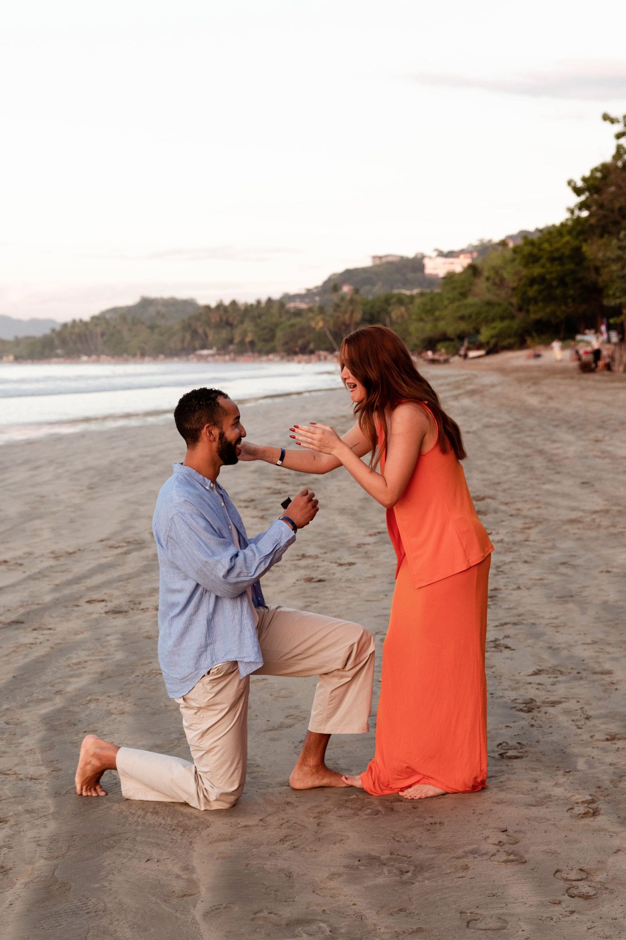 A surprise beach proposal moment showing a couple getting engaged on the sand at dusk.